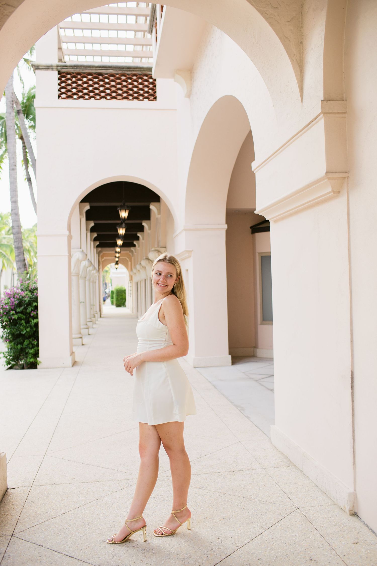Young blonde woman in white dress standing in arched corridor of upscale resort, smiling at camera.