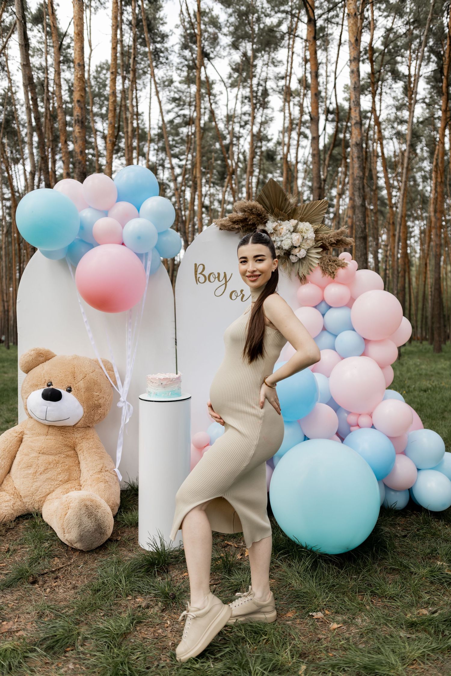 Gender reveal party setup with pink and blue balloons, white backdrop, and teddy bear in forest setting.