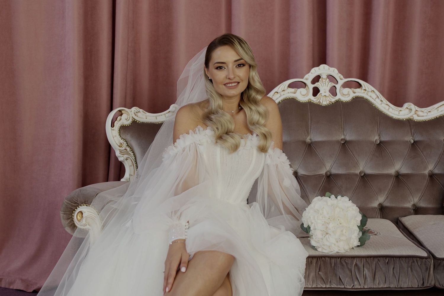 Smiling bride in white corset dress and veil seated on elegant tufted sofa with bouquet of white flowers.