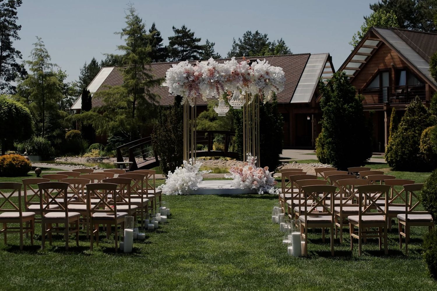 Elegant outdoor wedding ceremony setup with floral arch, wooden chairs, and candles on a lush green lawn.