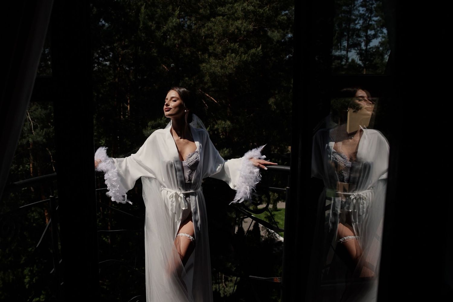 Woman in white sheer robe reflected in glass door surrounded by dark forest trees in artistic photography.