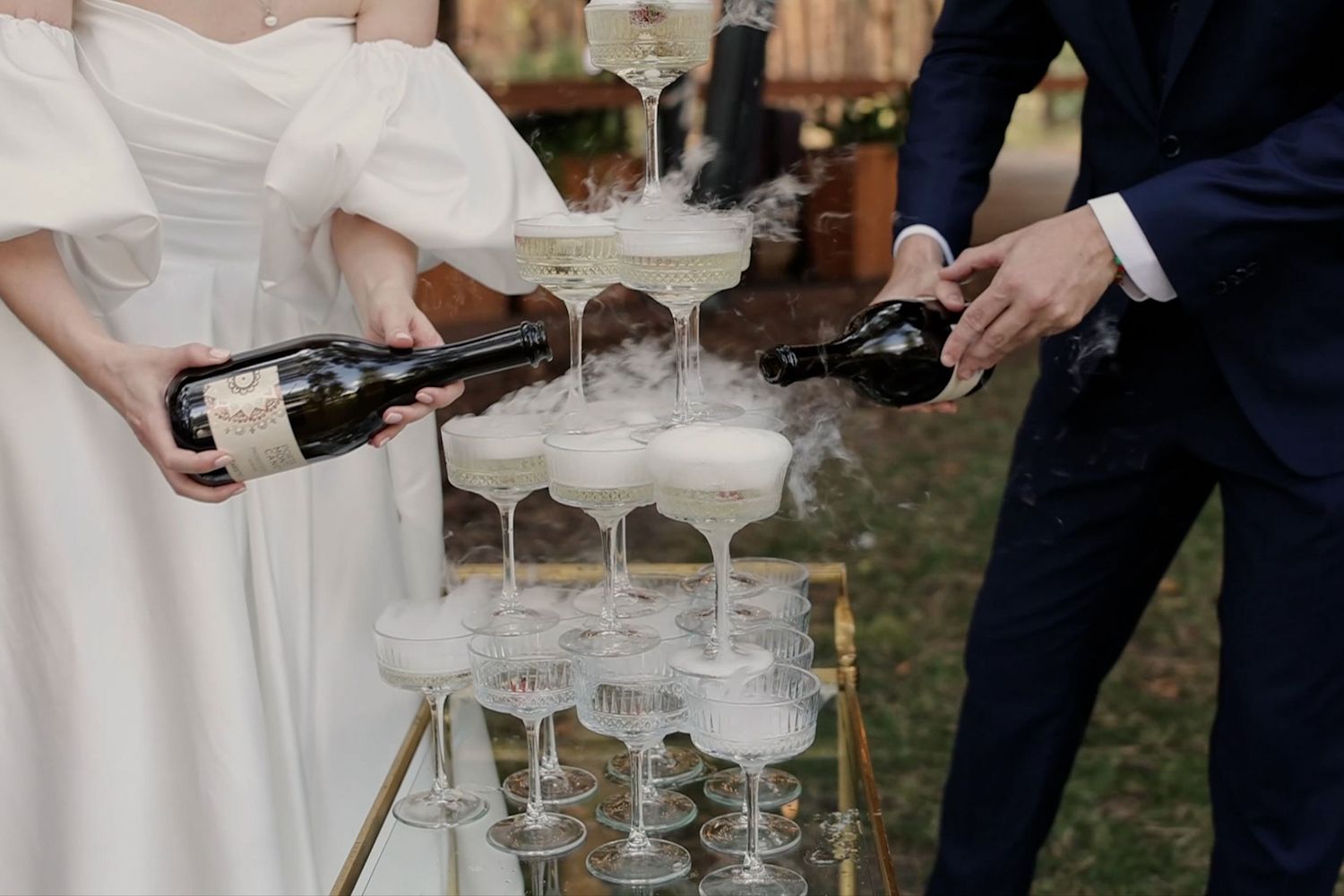 Bride and groom pouring champagne into a elegant tiered tower of coupe glasses with misty dry ice effect.