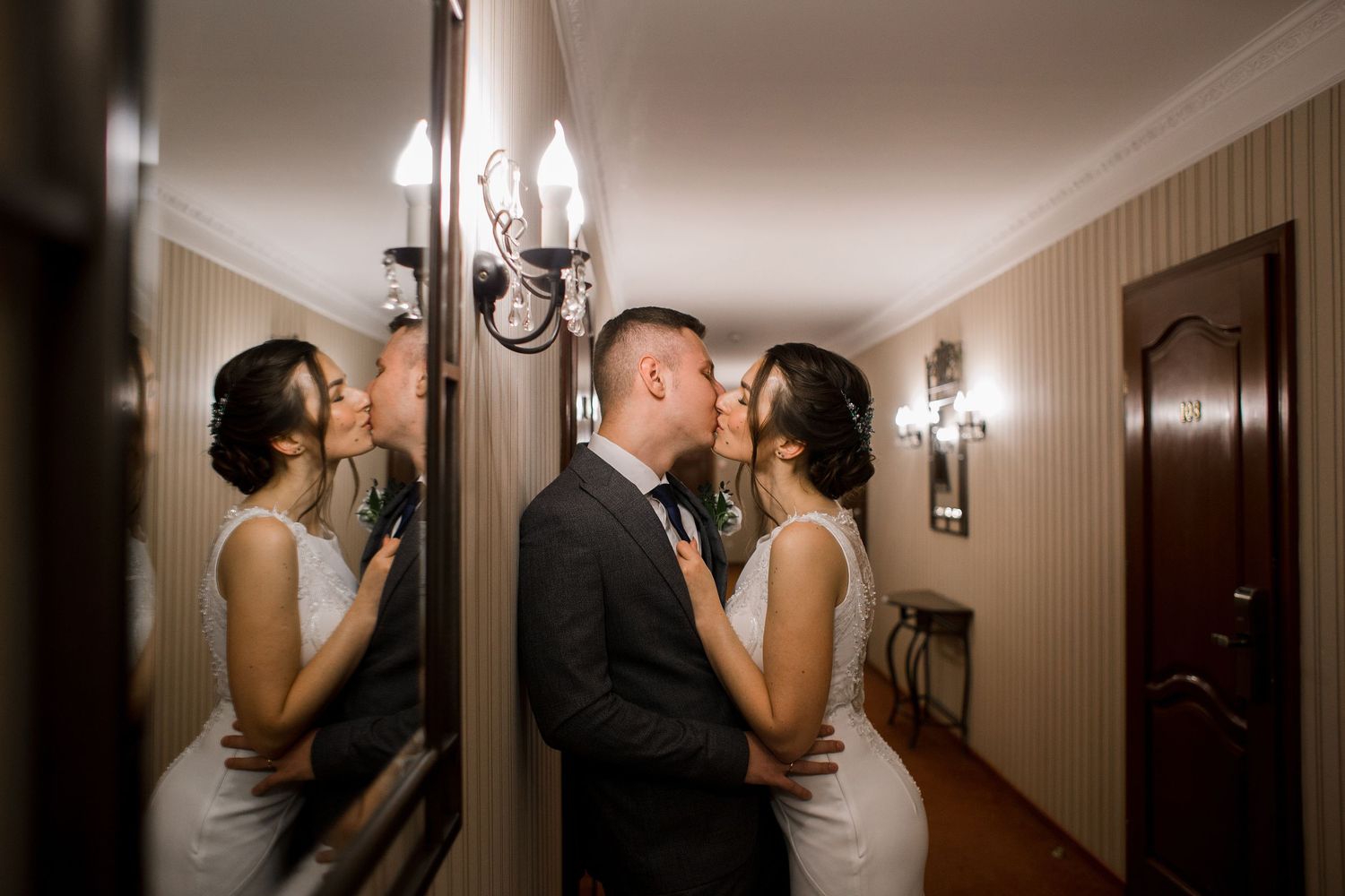 Bride and groom sharing a romantic kiss in a hallway, with their reflection visible in a large mirror beside them.