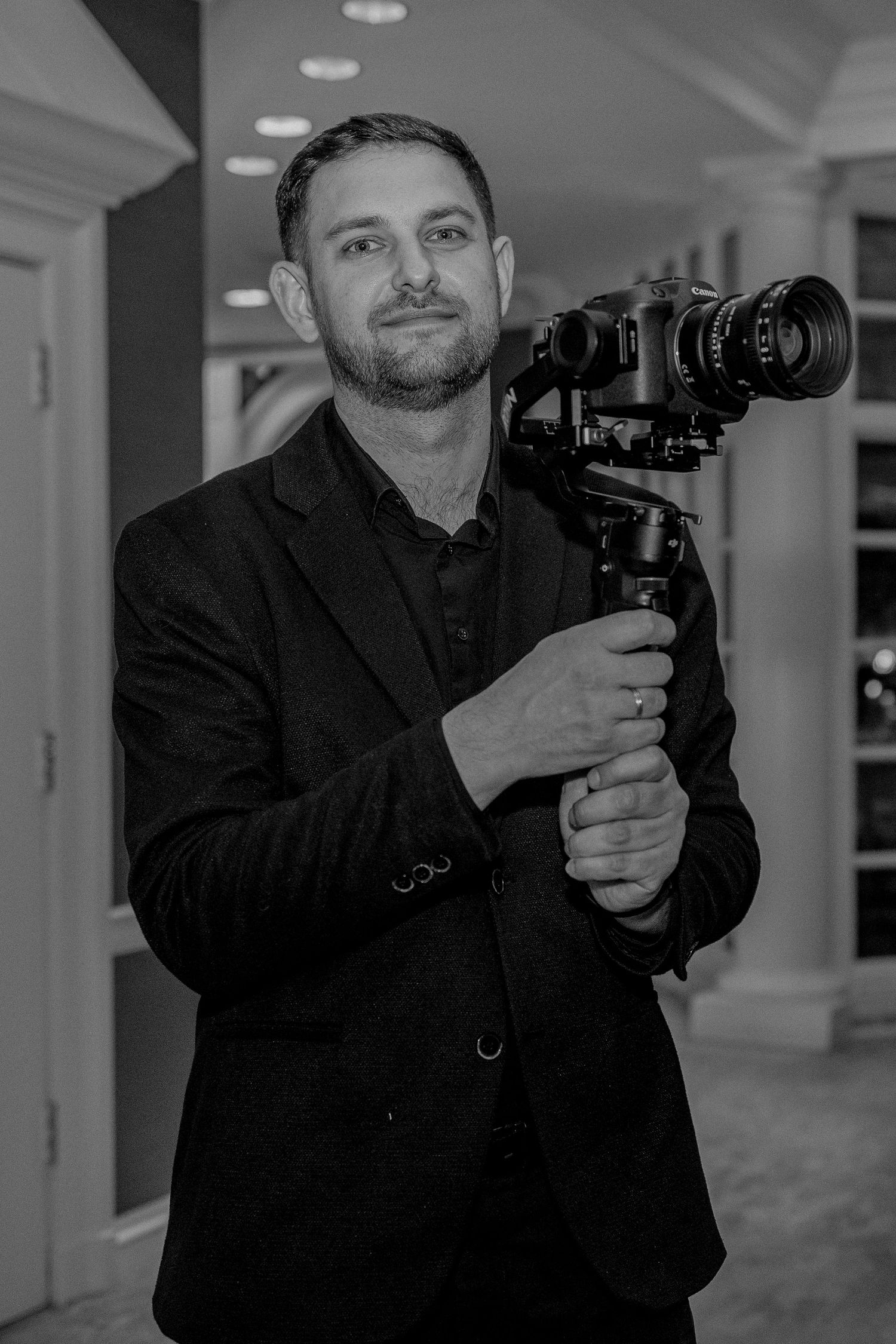 A professional videographer in a black suit holds a camera gimbal stabilizer in a black and white portrait photo.