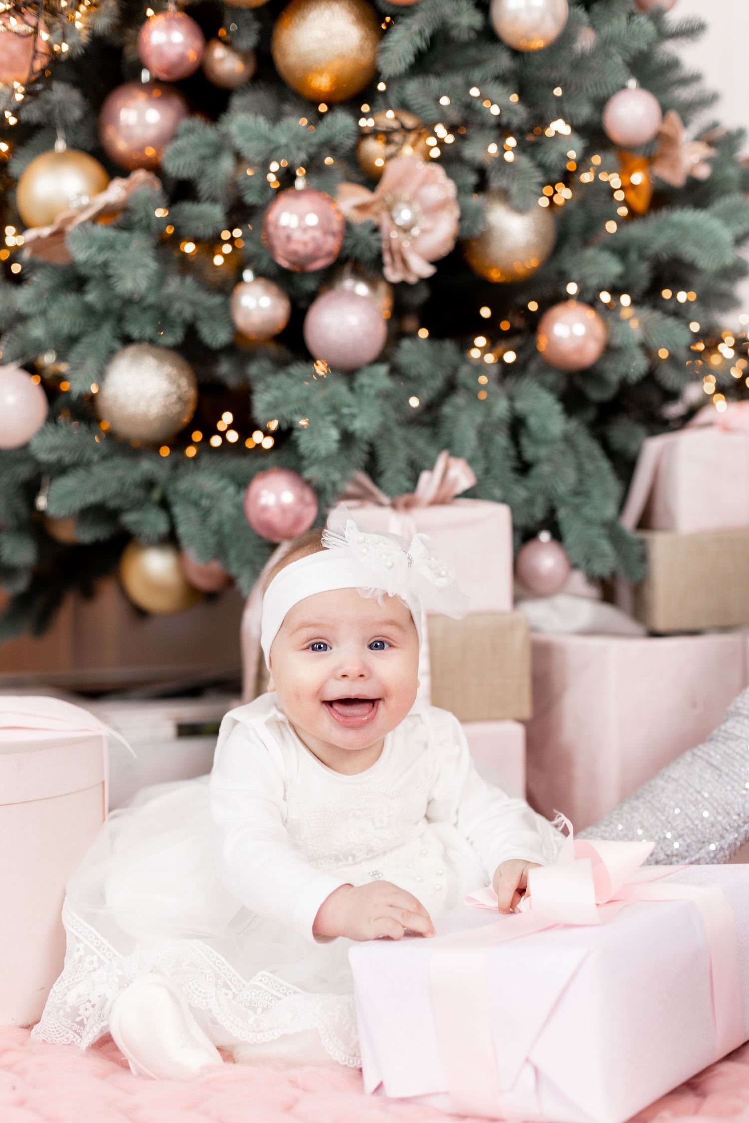 Baby in white dress lying among pink presents near decorated Christmas tree.
