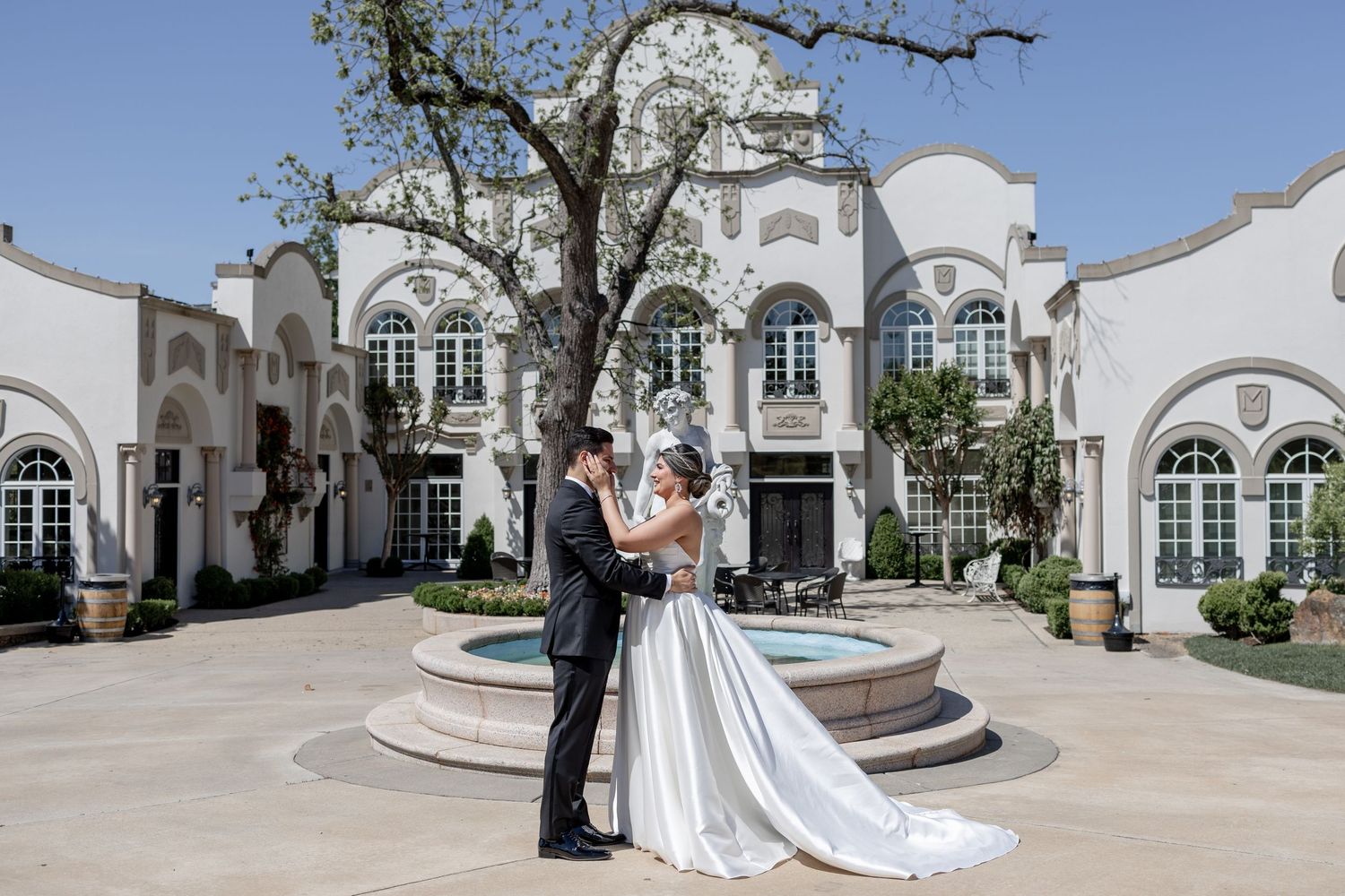 Bride and groom embrace by fountain outside elegant white wedding venue with arched windows and lush trees.