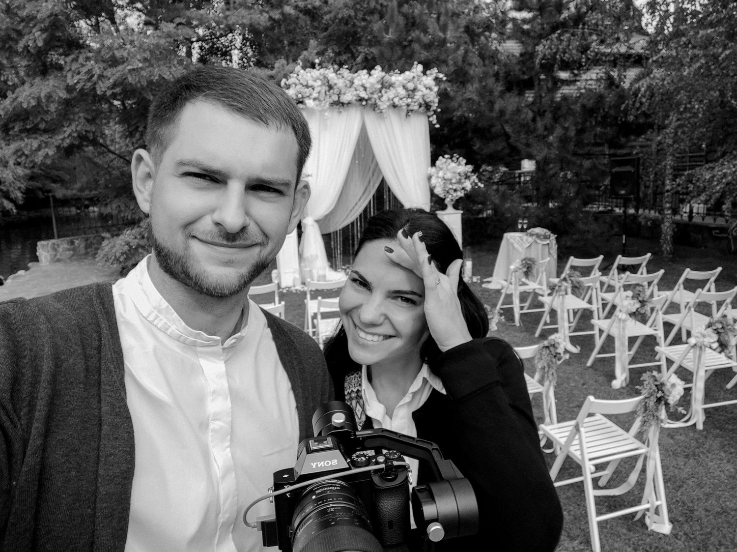 Black and white selfie of a smiling couple at an outdoor wedding venue with floral arch and white chairs.