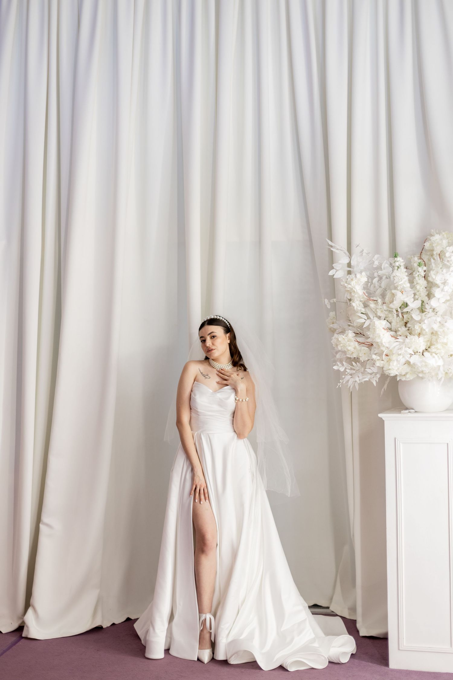 Elegant bride in a white satin gown with a thigh-high slit, posing near white curtains and floral arrangement.
