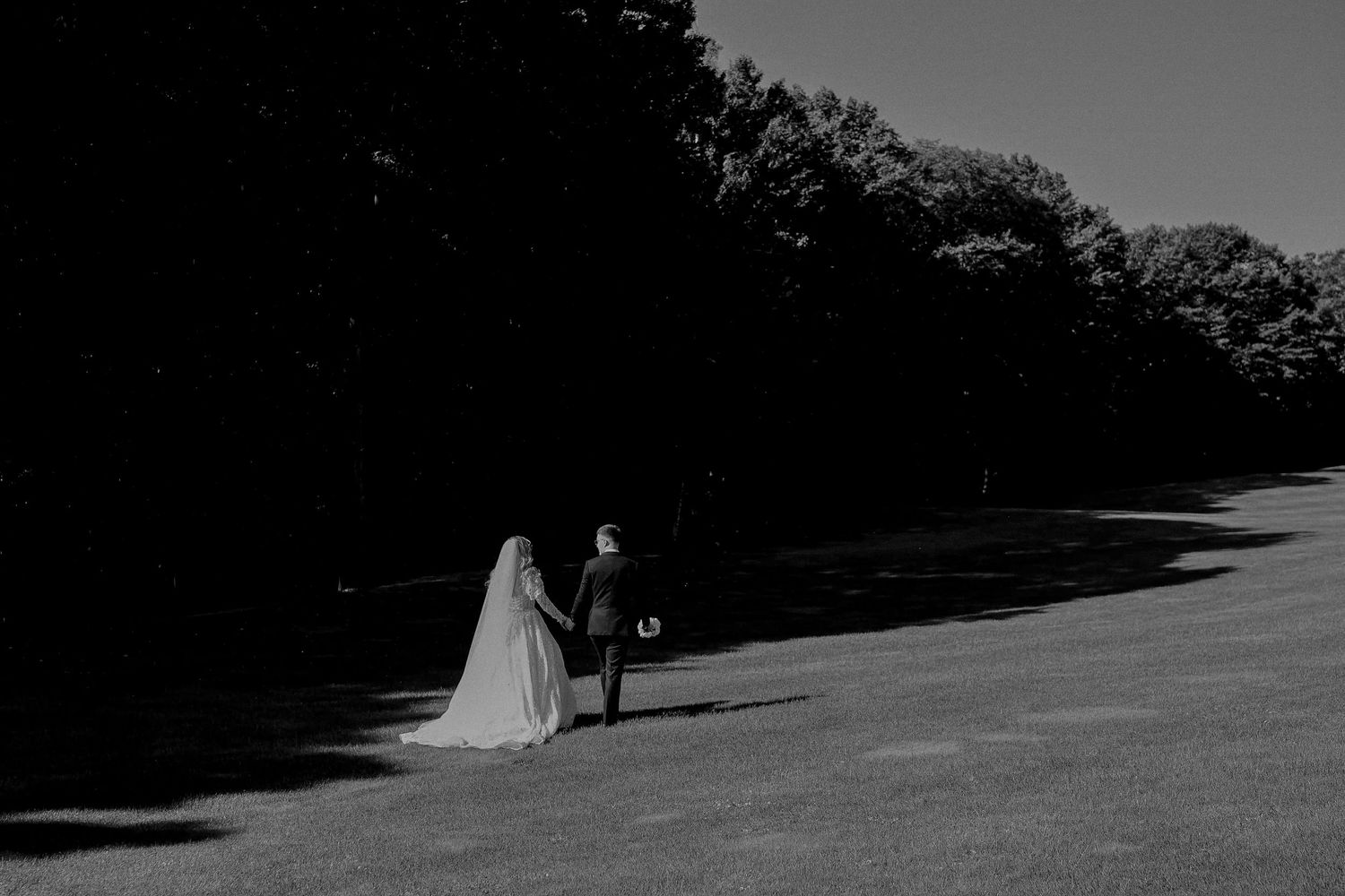 Black and white wedding photo of bride and groom walking hand in hand across a sunlit lawn surrounded by trees.