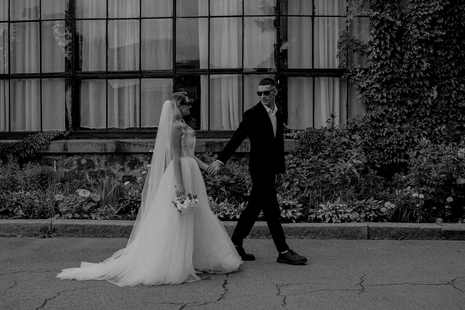 Black and white photo of a bride and groom walking hand in hand outside a building covered in ivy.