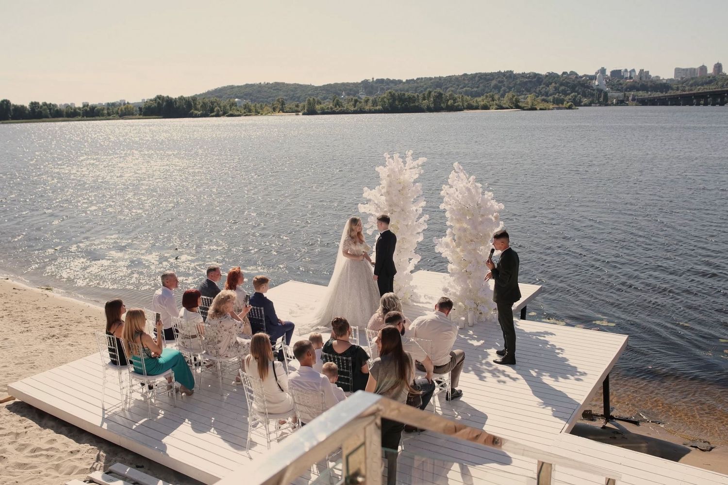 Outdoor wedding ceremony on a white dock by a lake, with white floral arrangements and seated guests on a sunny day.