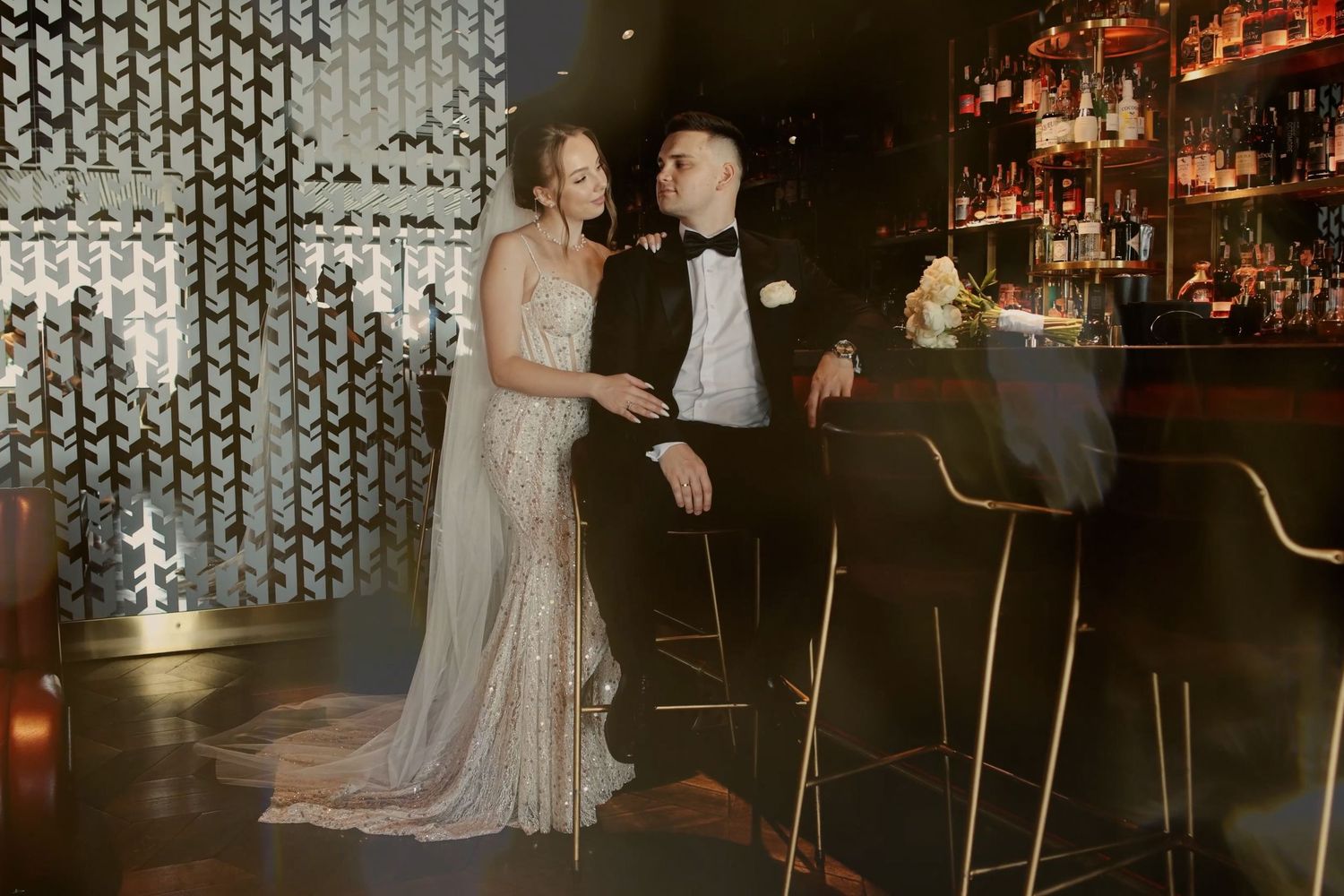 Bride and groom share a romantic moment at a dimly lit bar, bride in a sequined gown, groom in a black tuxedo.