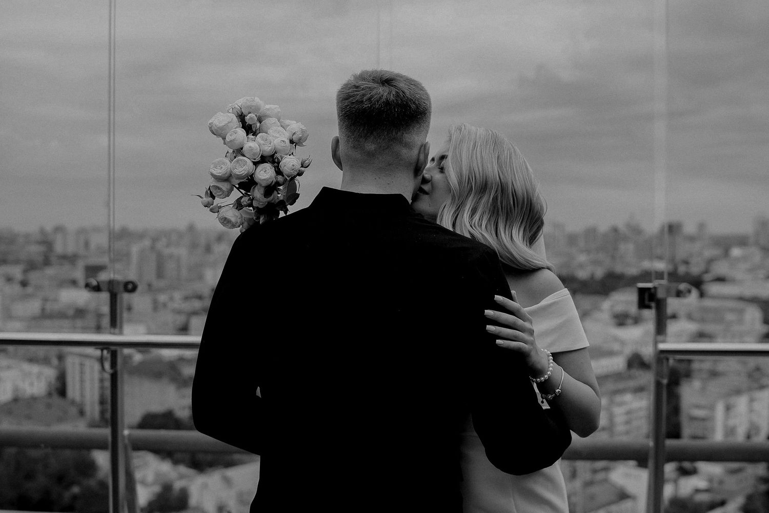 Black and white photo of a couple embracing on a rooftop, man holding bouquet, city skyline in background.