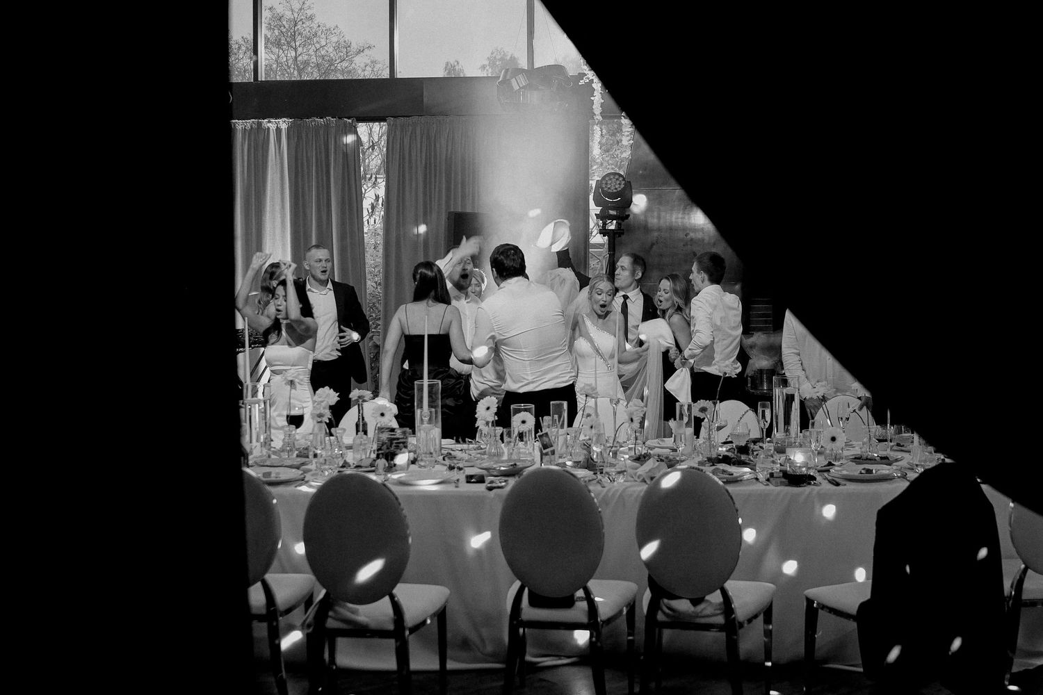 Black and white wedding reception photo showing guests gathered around a elegantly set head table with candles.