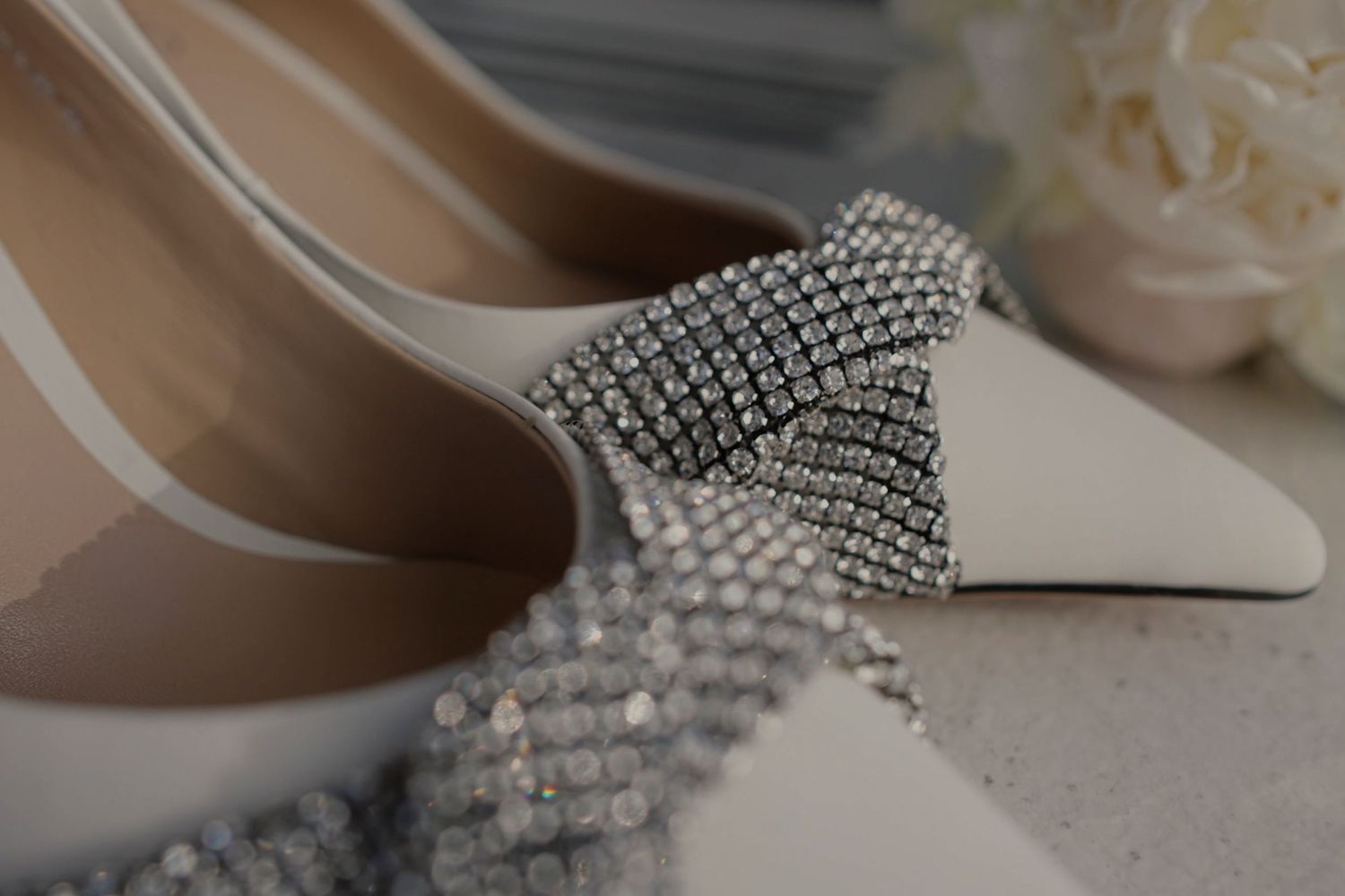 Elegant white bridal heels adorned with sparkling crystal rhinestone bow embellishments on a soft background.