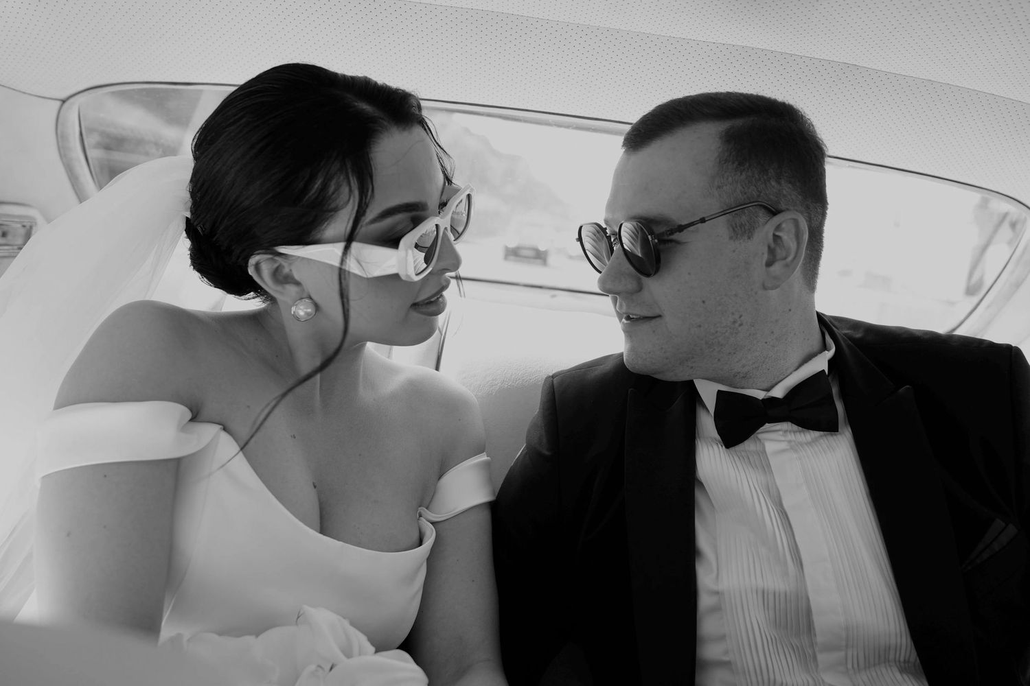 Black and white photo of a bride and groom in sunglasses sharing a romantic moment inside a car.