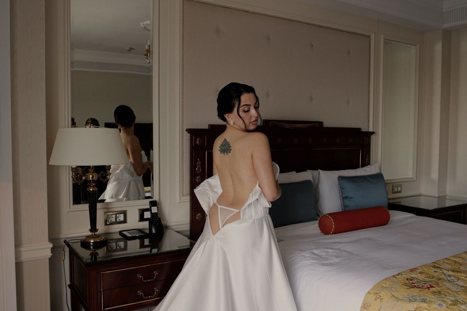 Bride in elegant backless white wedding gown with tattoo, standing in luxury hotel room beside bed and mirror.