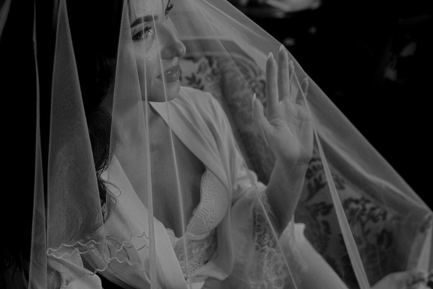 Black and white photo of a bride veiled in sheer fabric, hand raised, creating an ethereal wedding portrait.