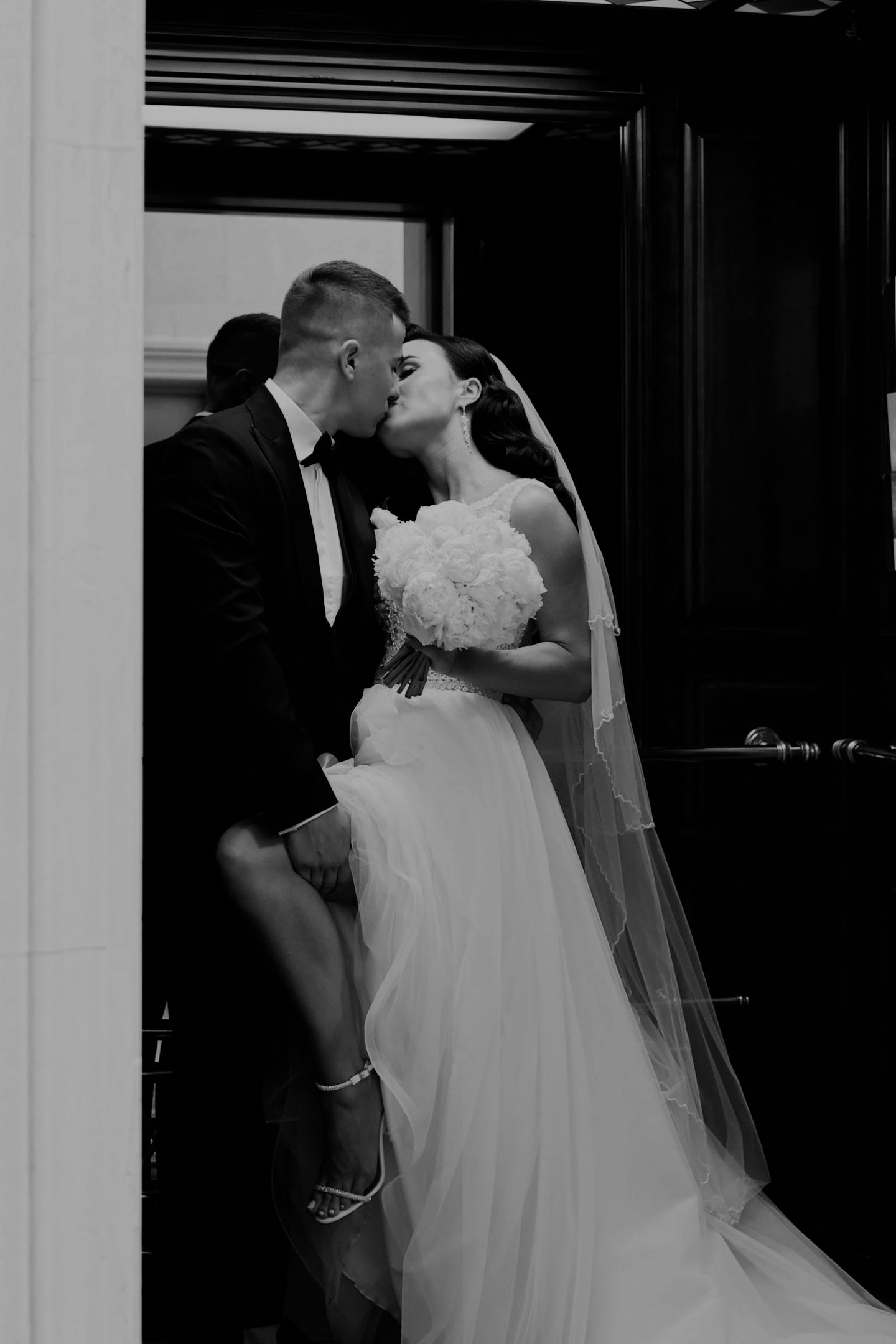 Black and white photo of a bride and groom sharing a kiss in a doorway, bride holding a bouquet.