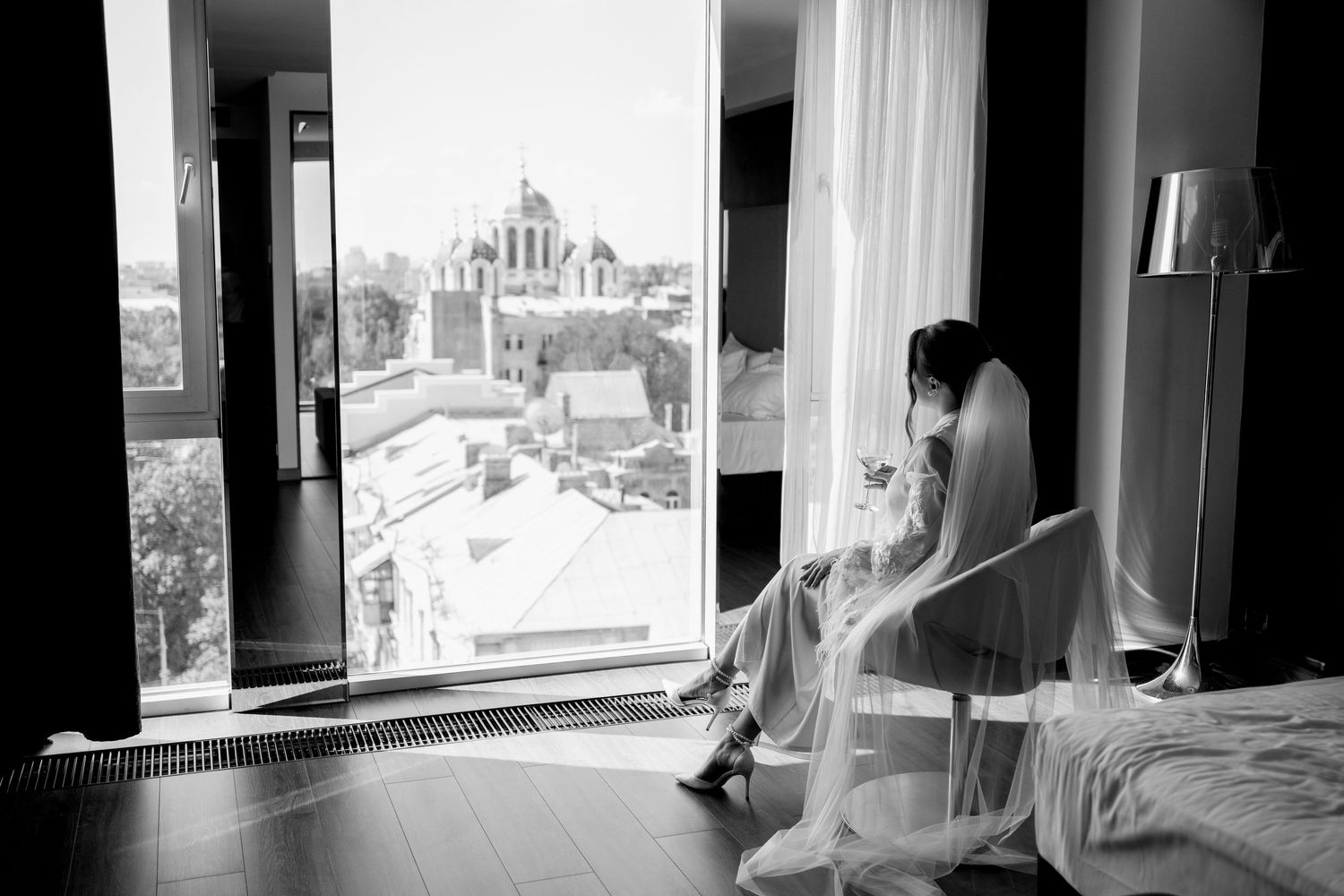 Bride in wedding dress sitting by large window with city skyline view, black and white photography.