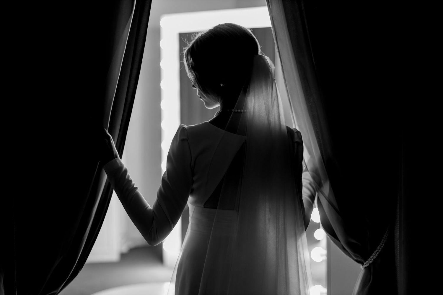 Black and white photo of a bride in a wedding dress and veil standing at a window between curtains.