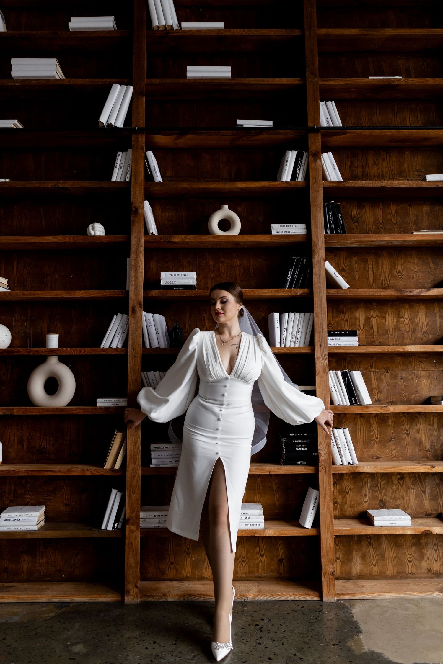 Elegant bride in white dress and veil poses gracefully against wooden bookshelves filled with books and decorative vases.