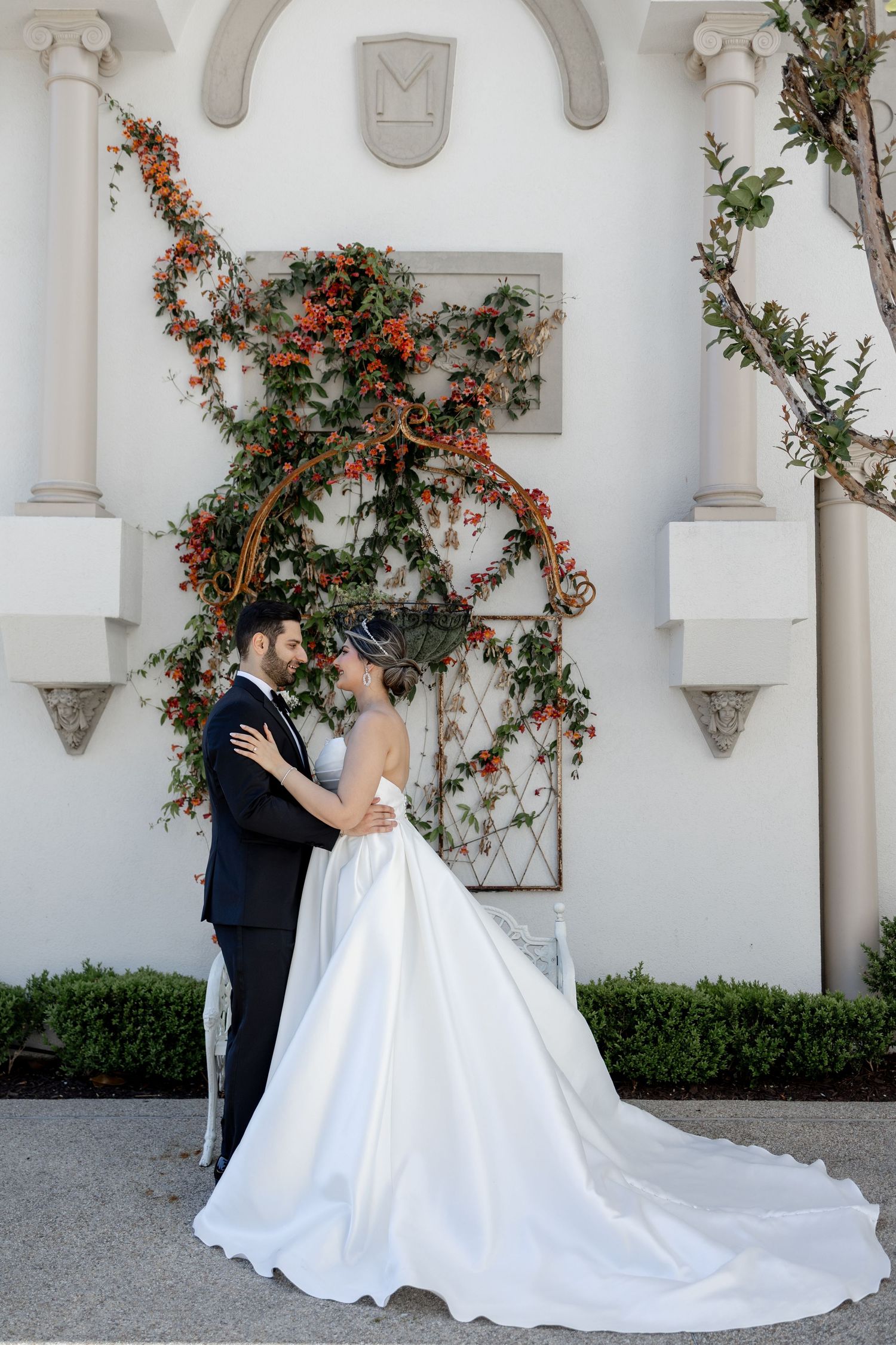 Bride and groom embracing before a white villa wall adorned with climbing vines and orange berries on their wedding day.