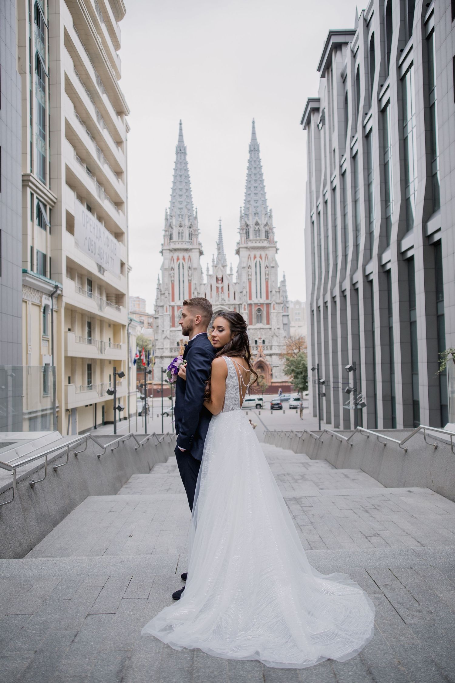 Bride and groom pose on steps with Gothic church towers in background during urban wedding photo session.
