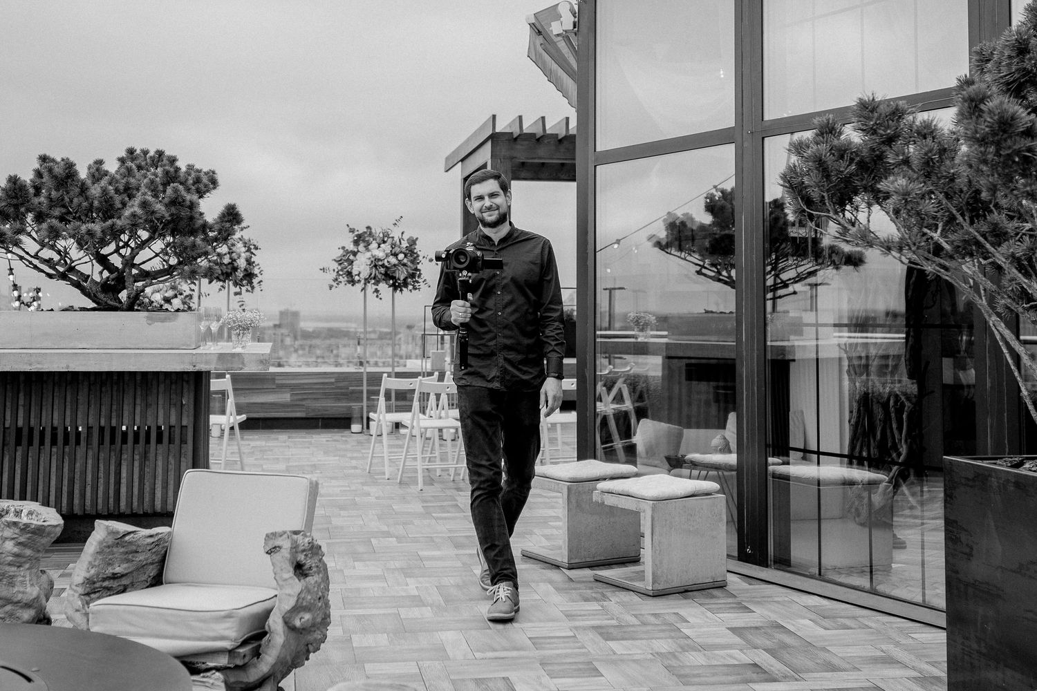 Person walking on rooftop terrace holding flowers, modern glass building backdrop, black and white photography.