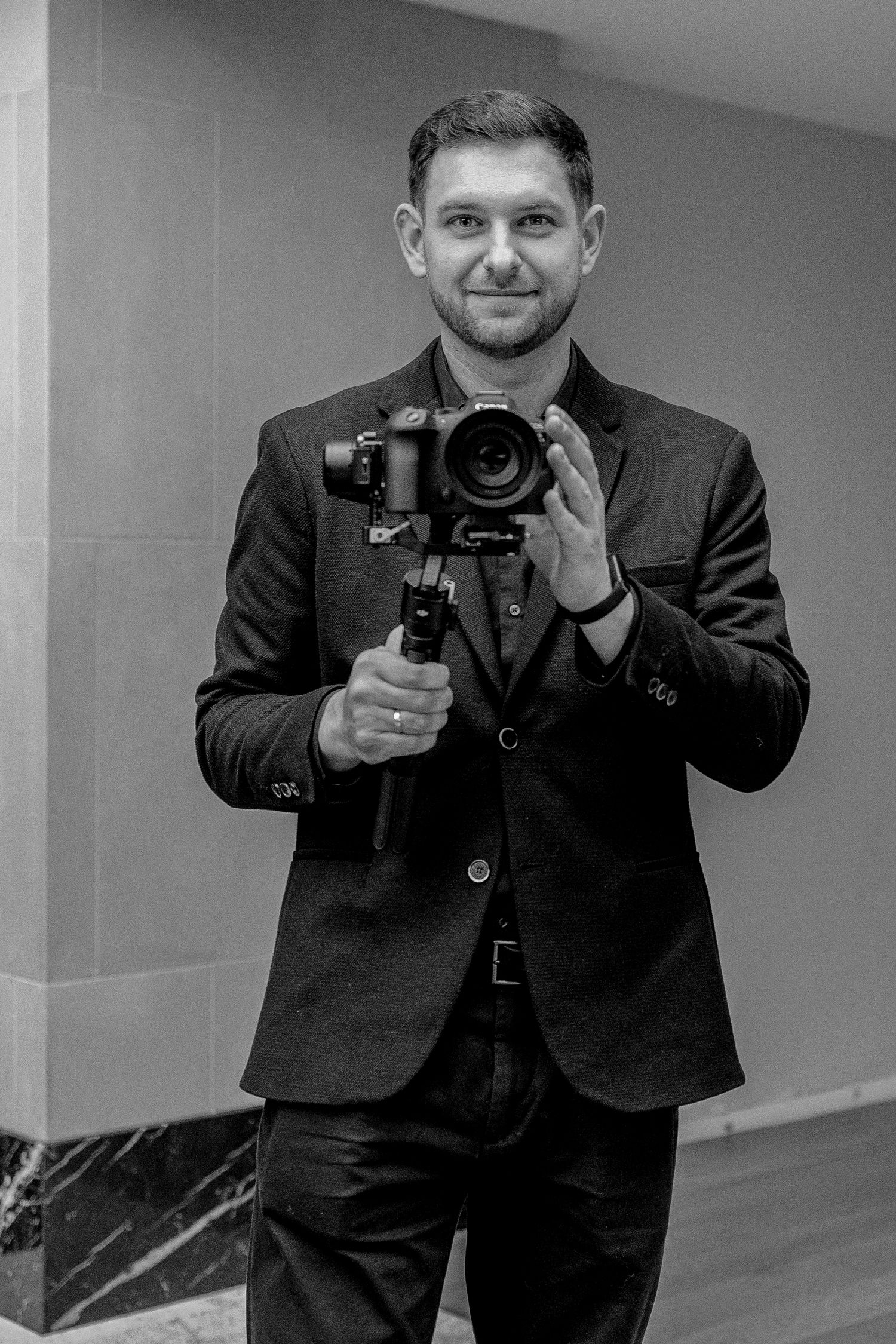 A smiling man in a black suit holds a professional camera on a gimbal stabilizer in a black and white portrait photo.