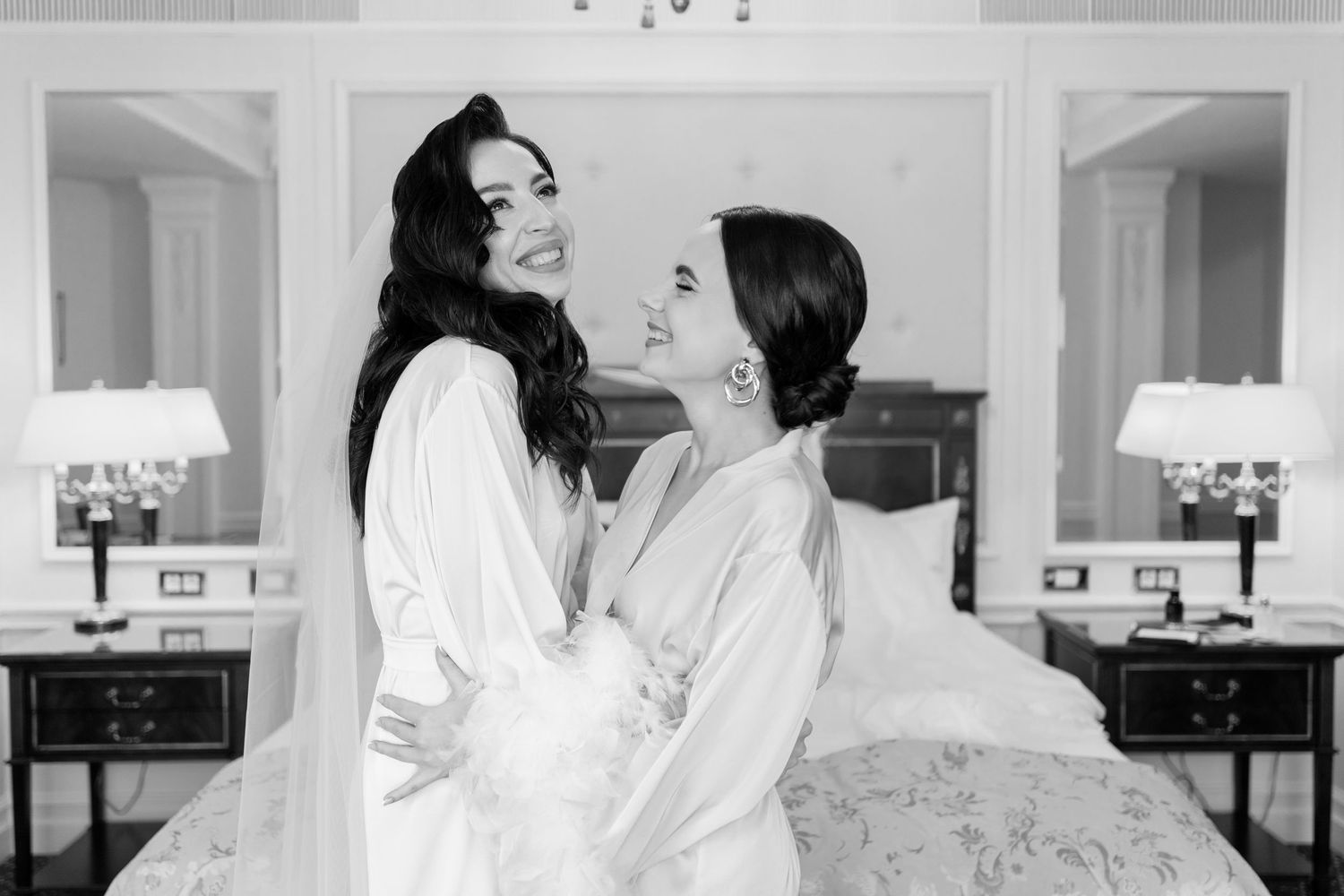 Two brides in white robes embrace and laugh together in an elegant hotel room on their wedding day.