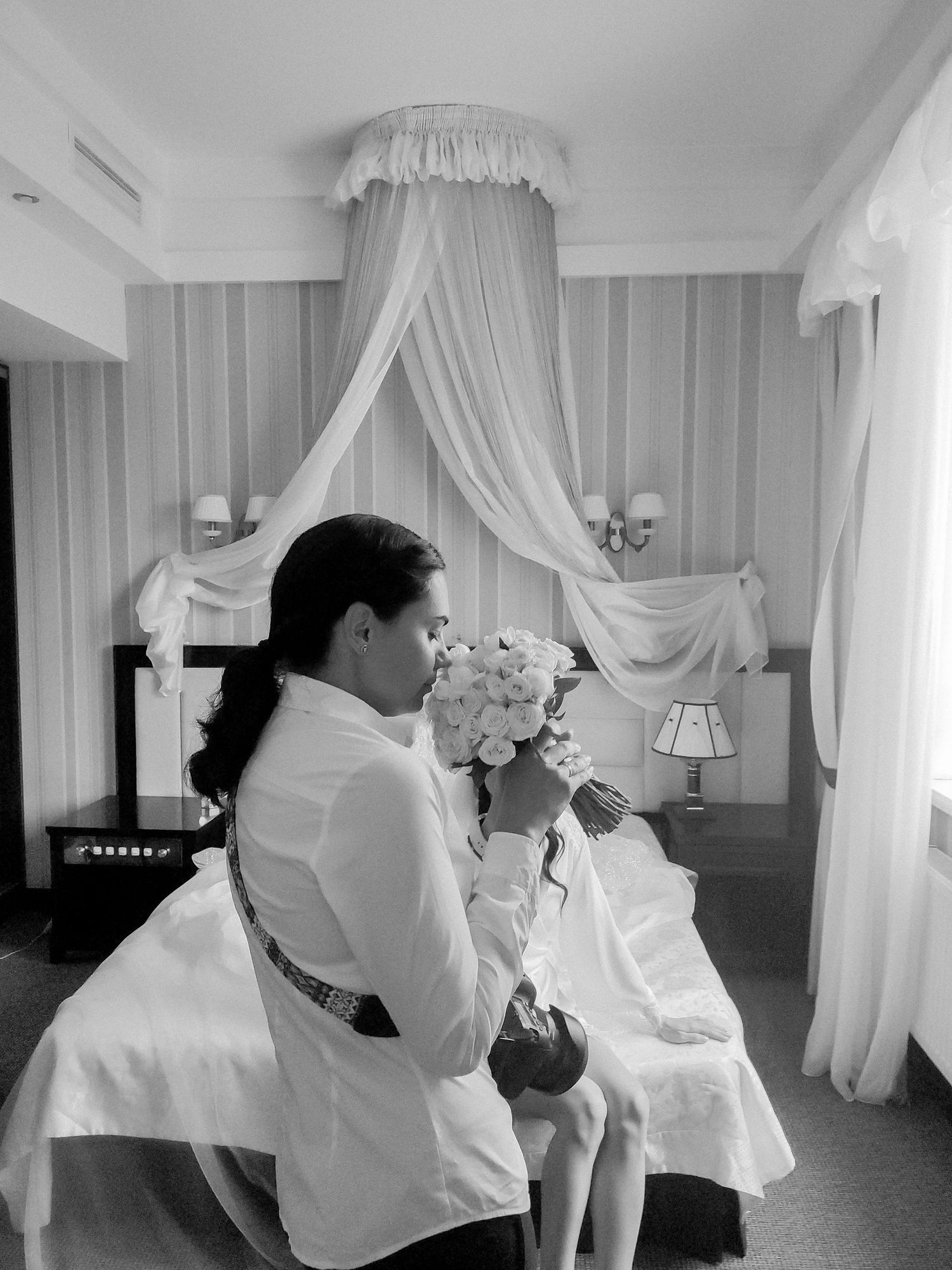 Black and white photo of a bride smelling her bouquet while sitting in an elegant hotel room with draped canopy.