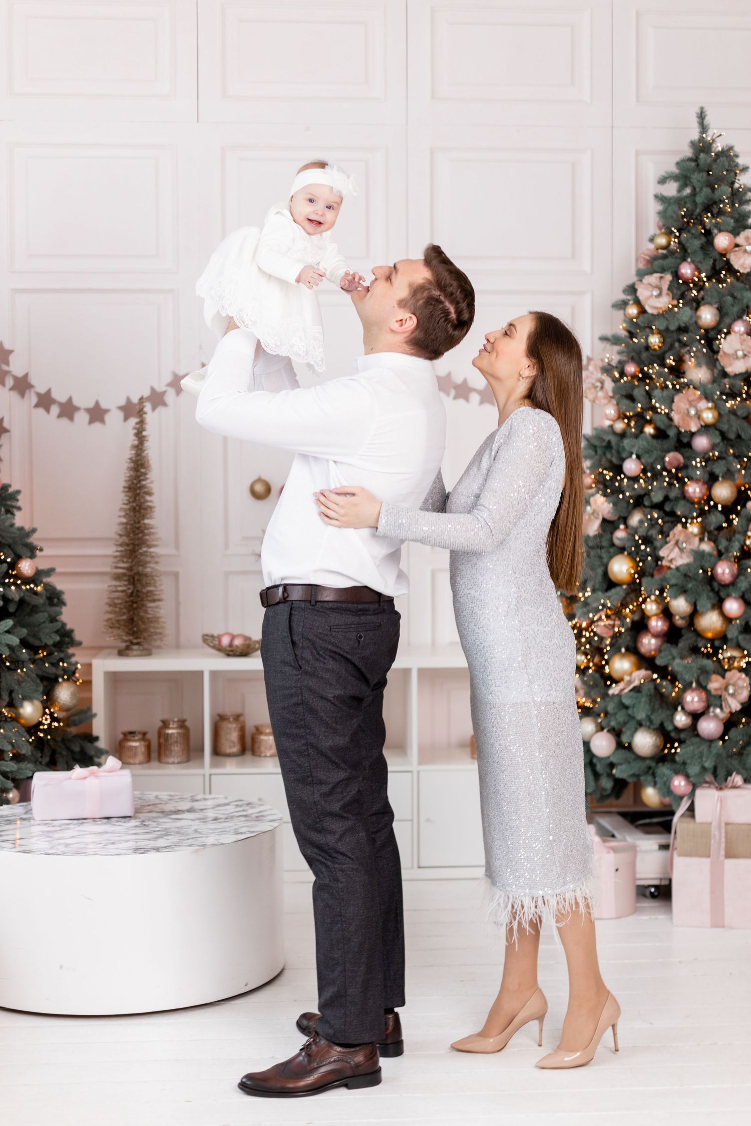 Family celebrating Christmas in front of decorated tree with baby in white dress.
