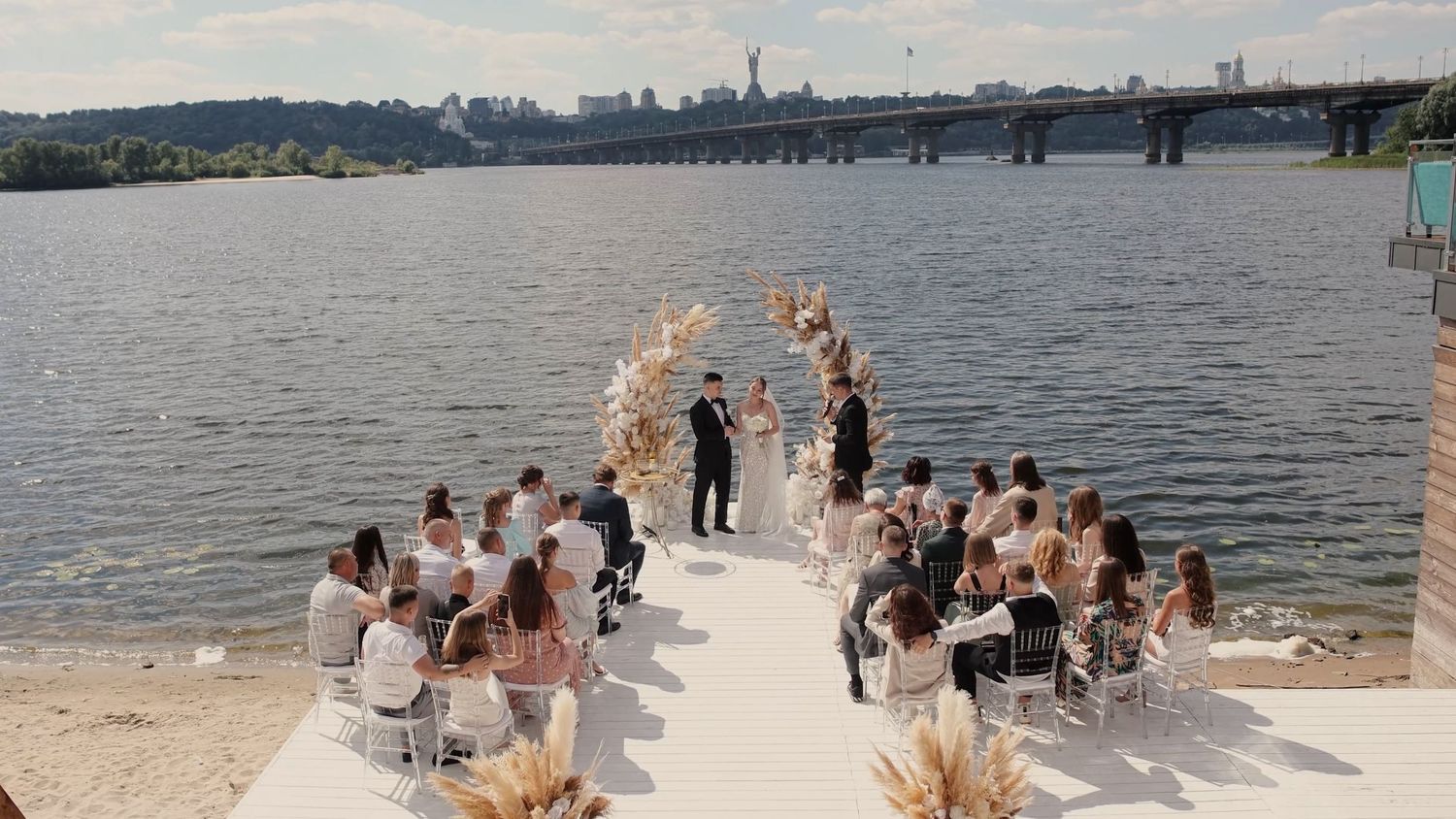 Outdoor wedding ceremony on a white pier by a river with floral arch, seated guests, and a city bridge backdrop.