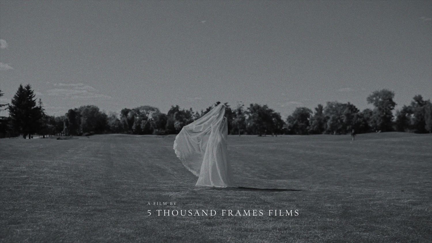 Black and white photo of a bride in flowing wedding dress standing alone in an open field by 5 Thousand Frames Films.
