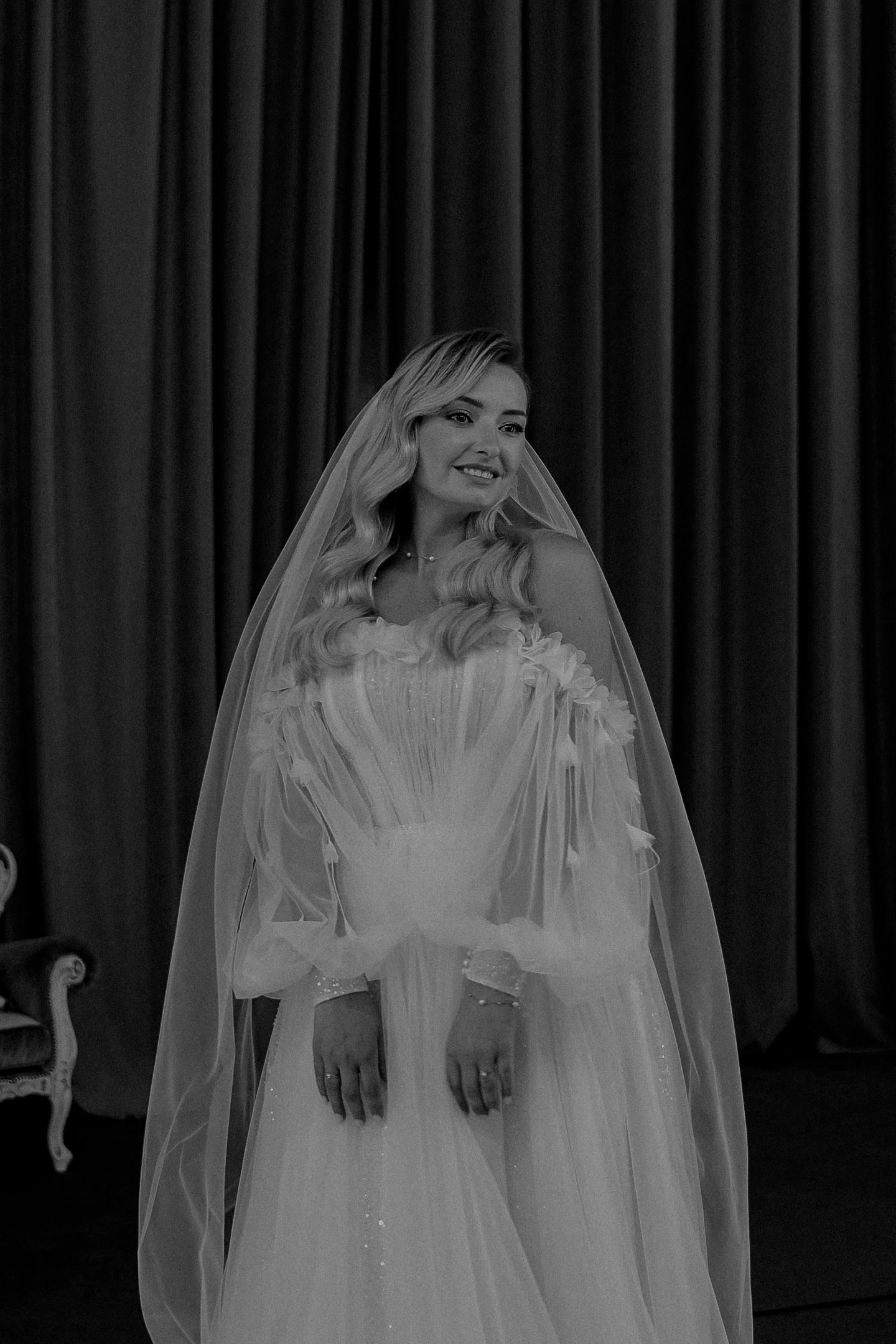 Smiling bride in a white tulle wedding dress and long veil poses elegantly in a black and white portrait photo.