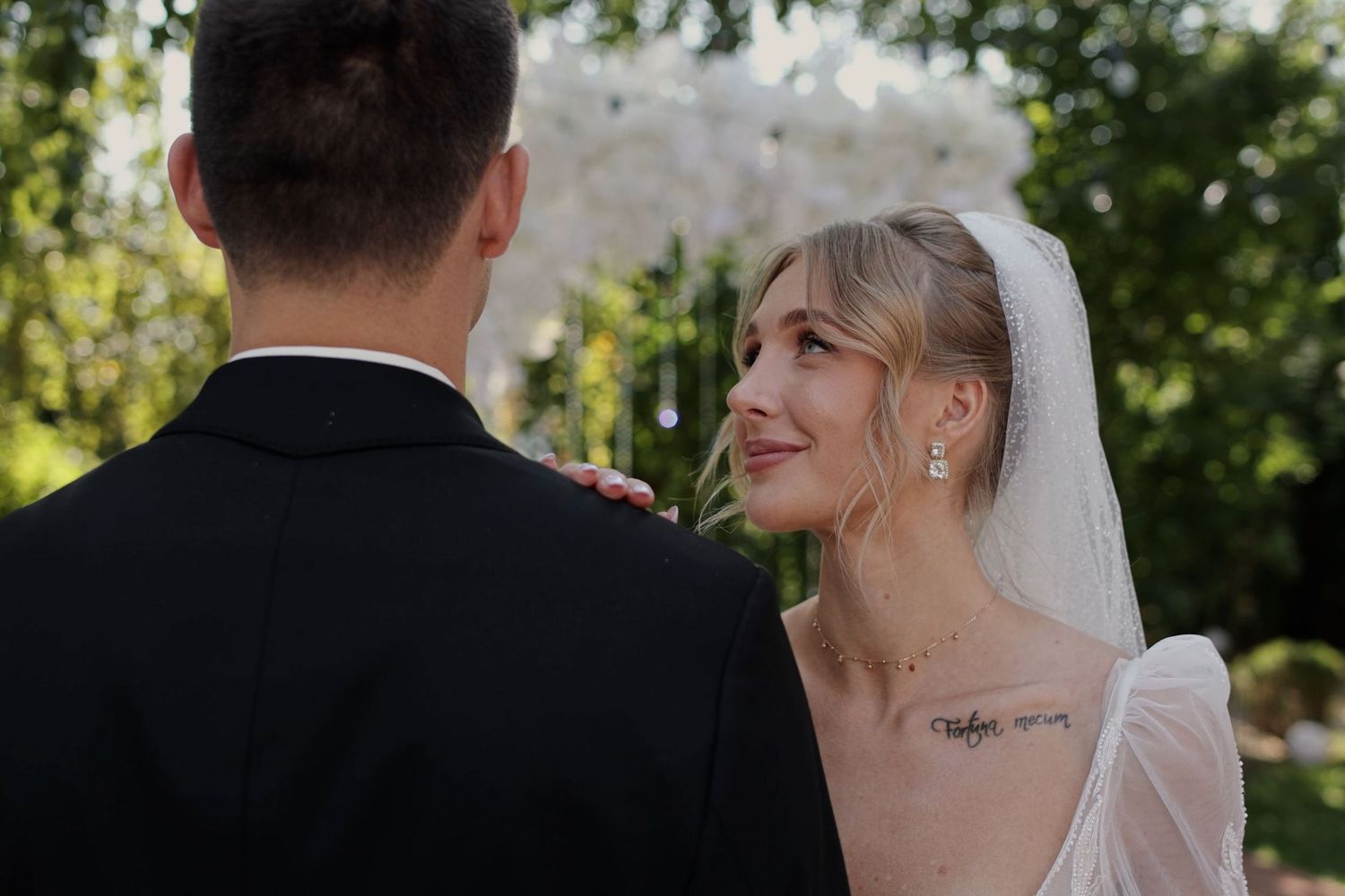 Bride in white veil and gown smiles at groom during outdoor wedding ceremony with floral arch backdrop.