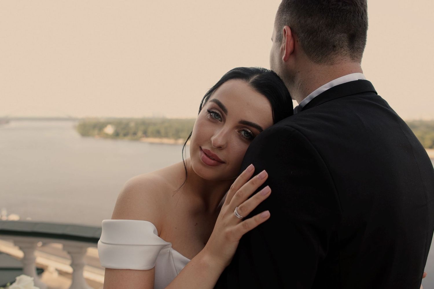 A bride in a white off-shoulder gown embraces her groom in a black suit outdoors near a waterfront at sunset.