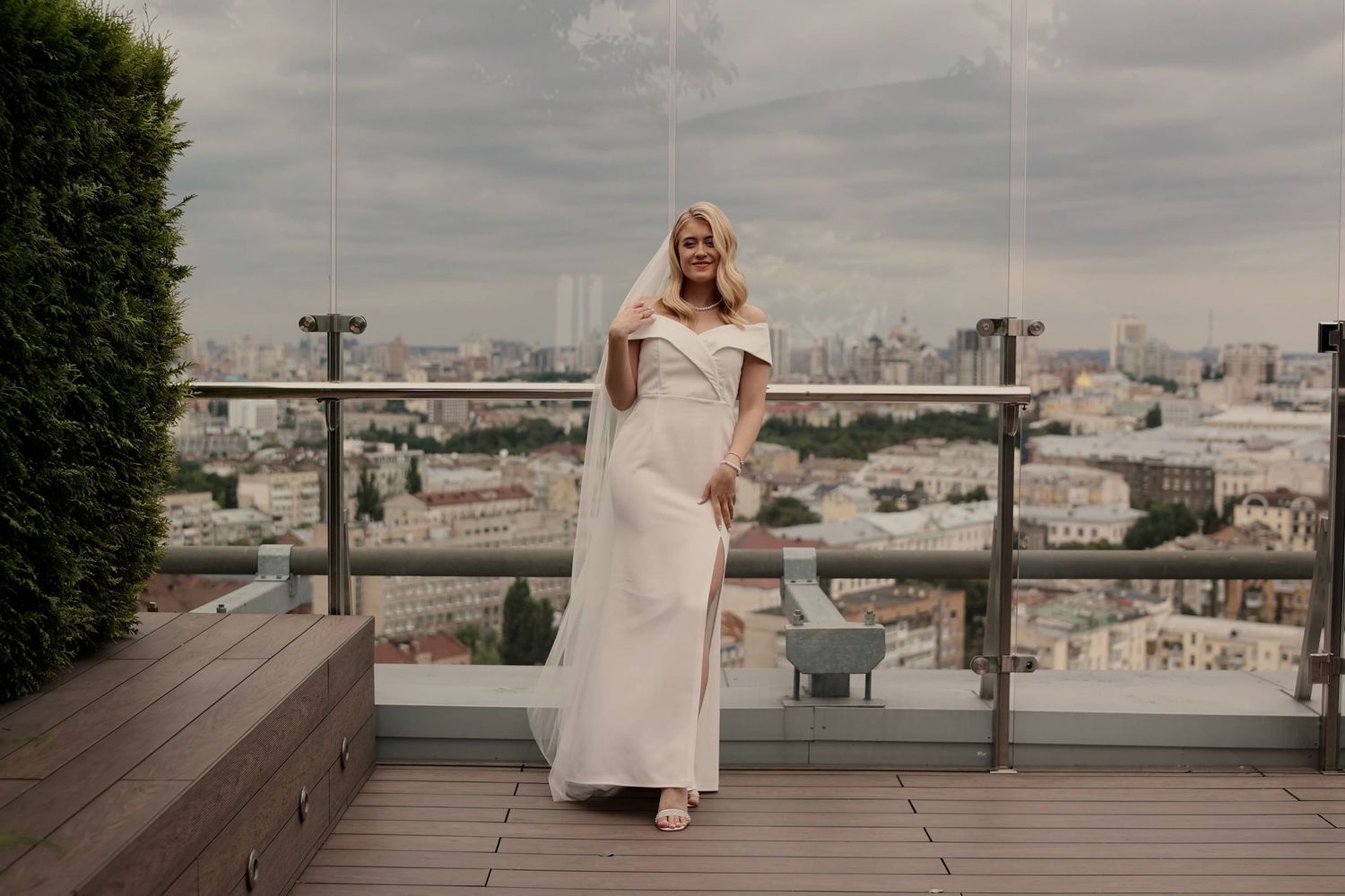 Blonde woman in elegant white off-shoulder gown poses on rooftop terrace with city skyline backdrop.