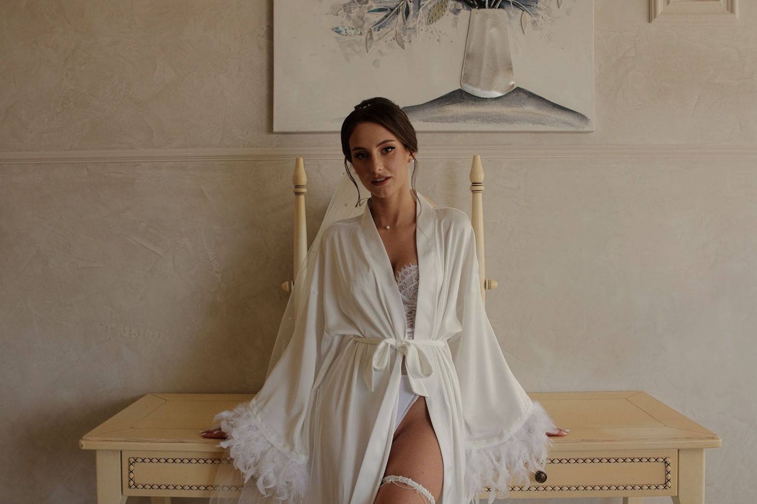 Woman in white bridal robe and lace lingerie sitting on dresser in elegant room with artwork.