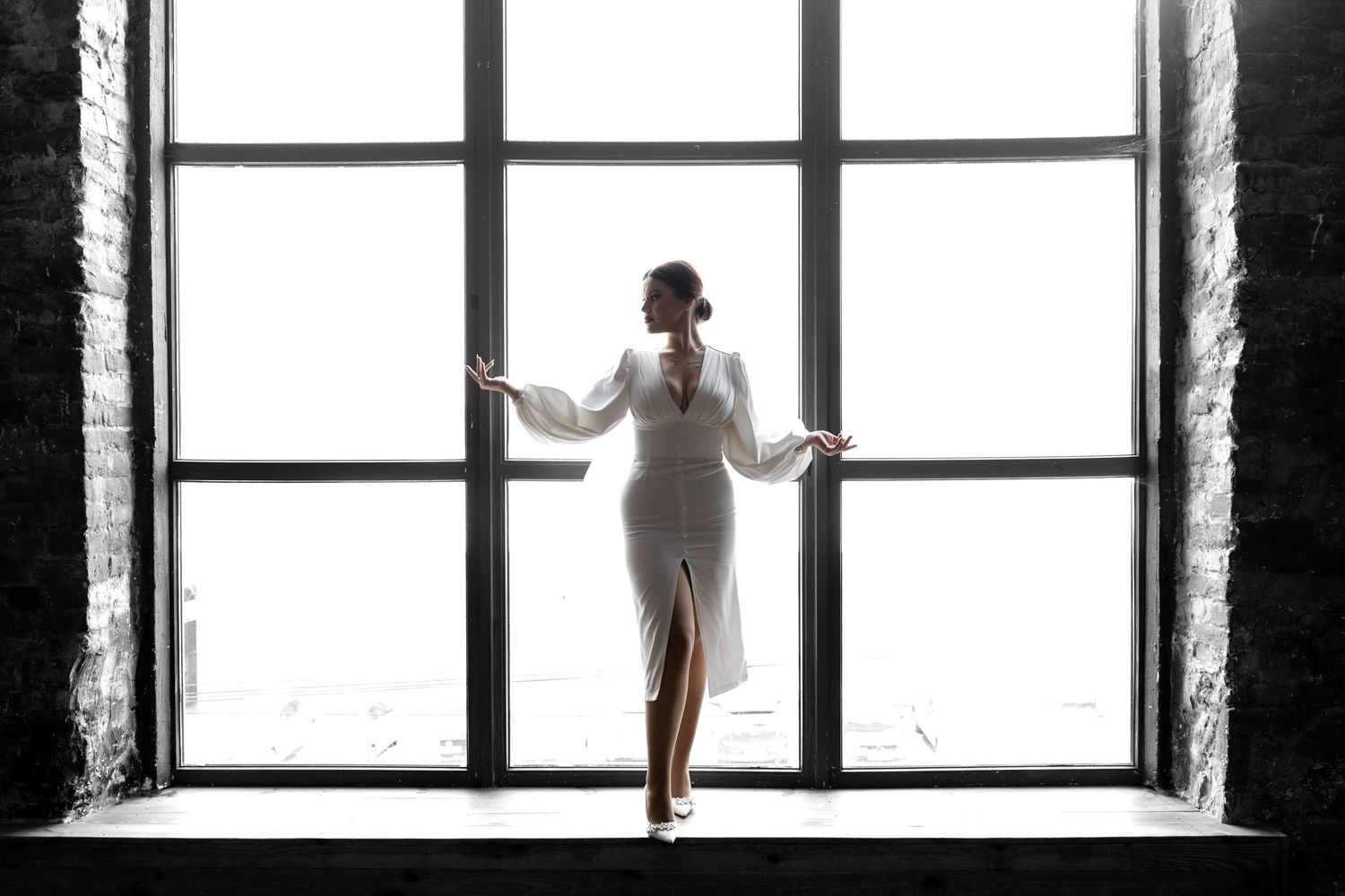 Woman in white dress standing before large window, silhouetted against bright light in elegant interior.
