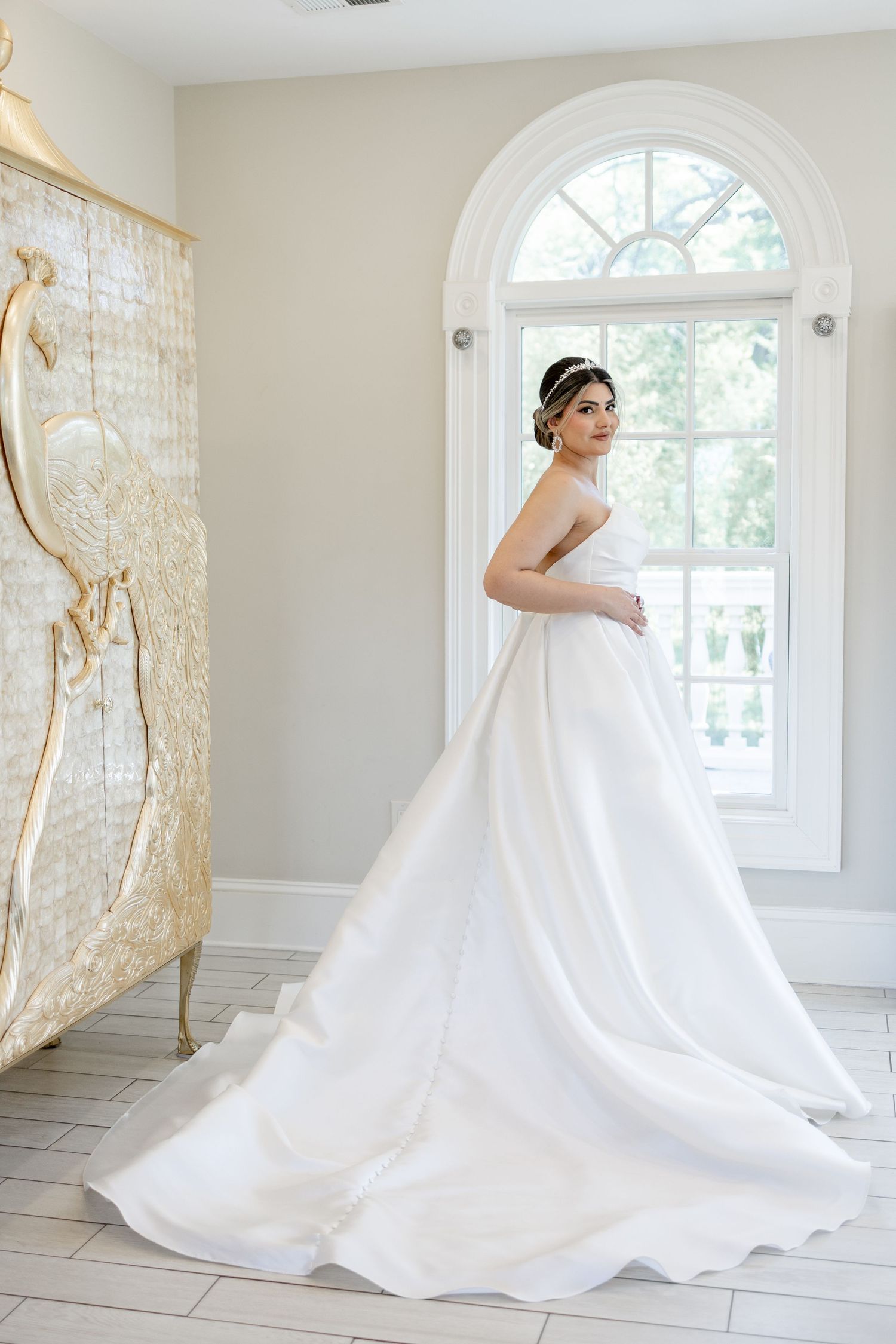 Bride in elegant white strapless ball gown with long train stands by arched window in bright room.