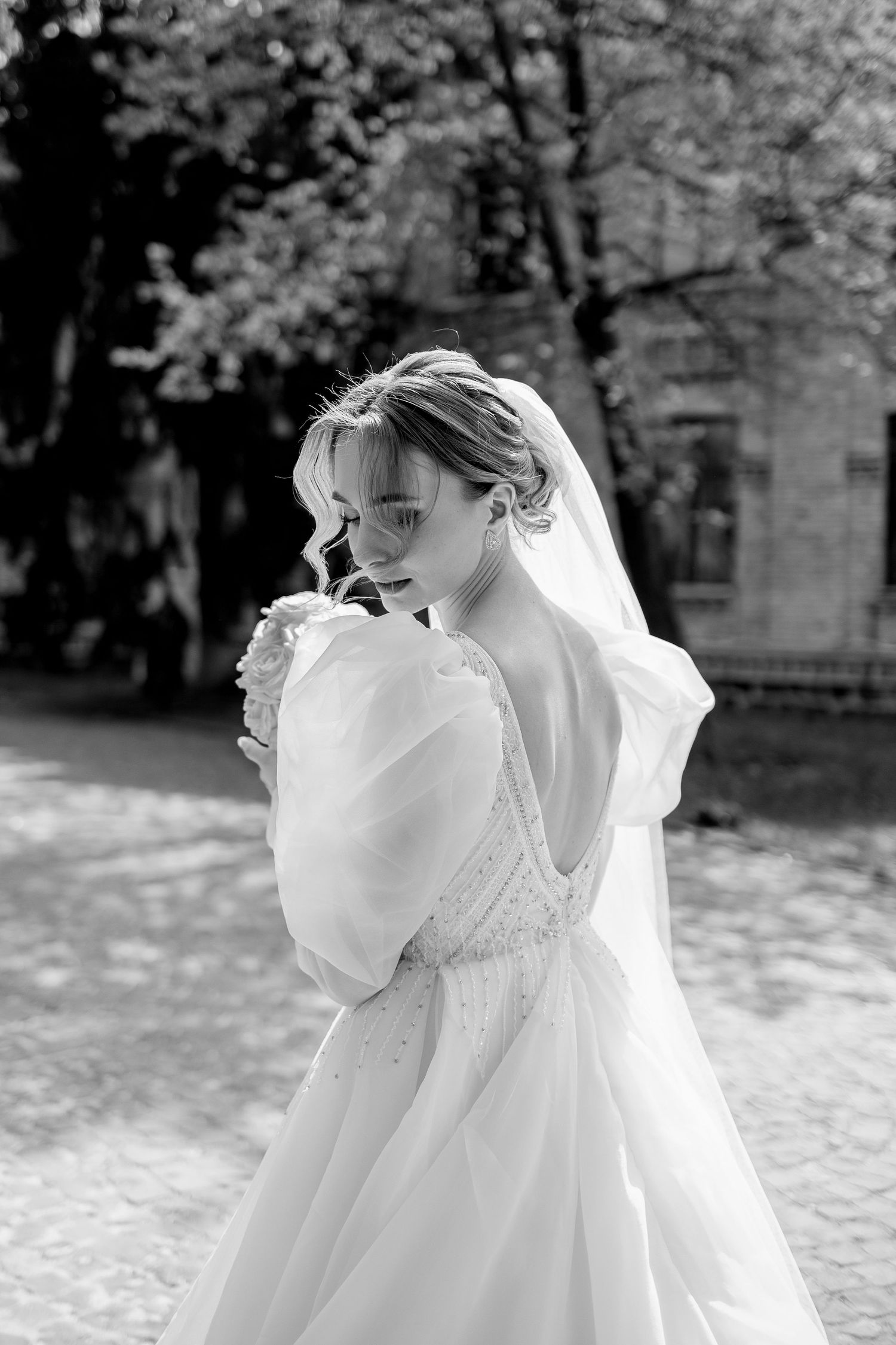 Black and white photo of a bride in a white gown with puff sleeves and veil, posing outdoors near trees.