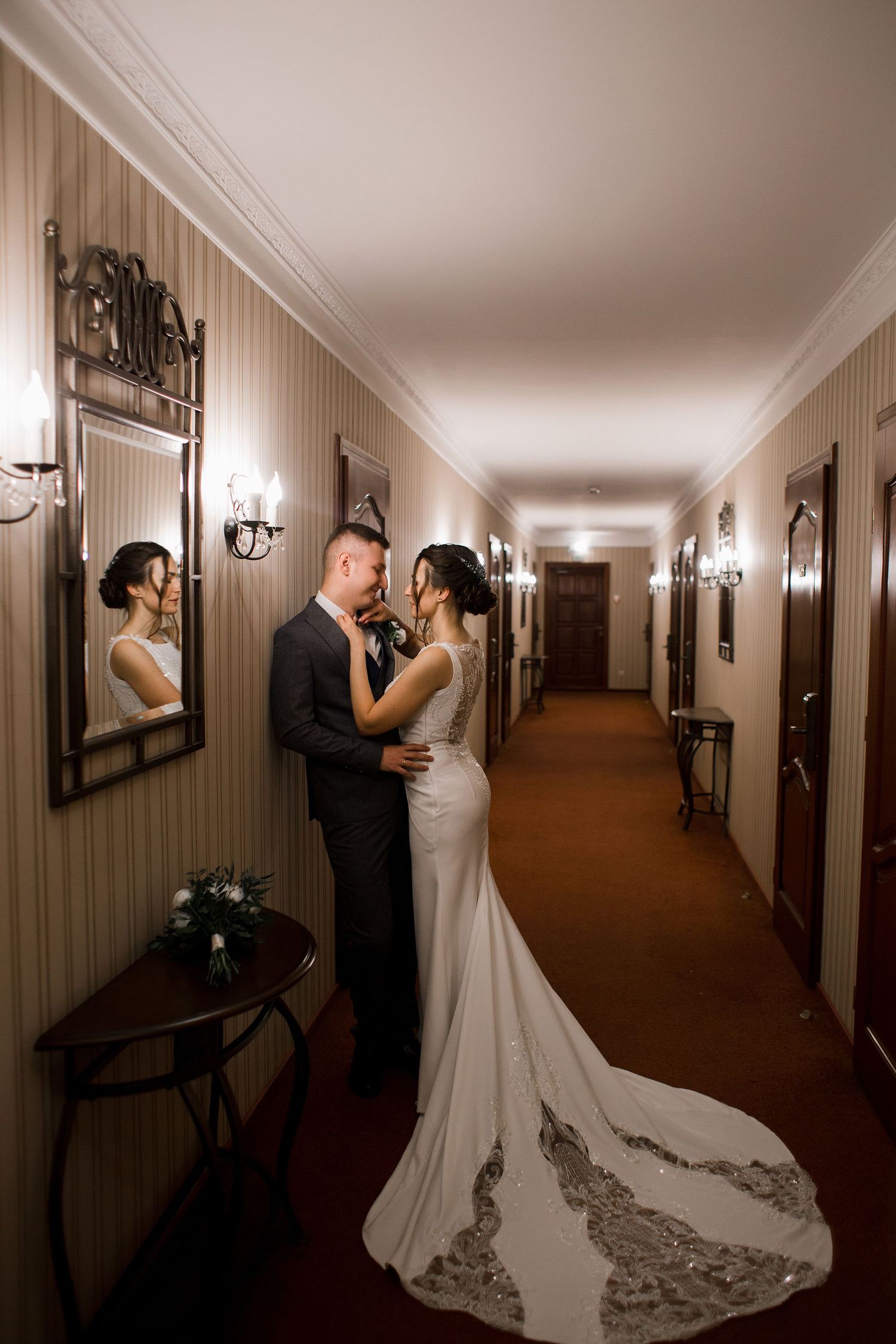 Bride and groom embracing in an elegant hotel corridor, with a mirror reflection, warm lighting, and a lace wedding gown train.