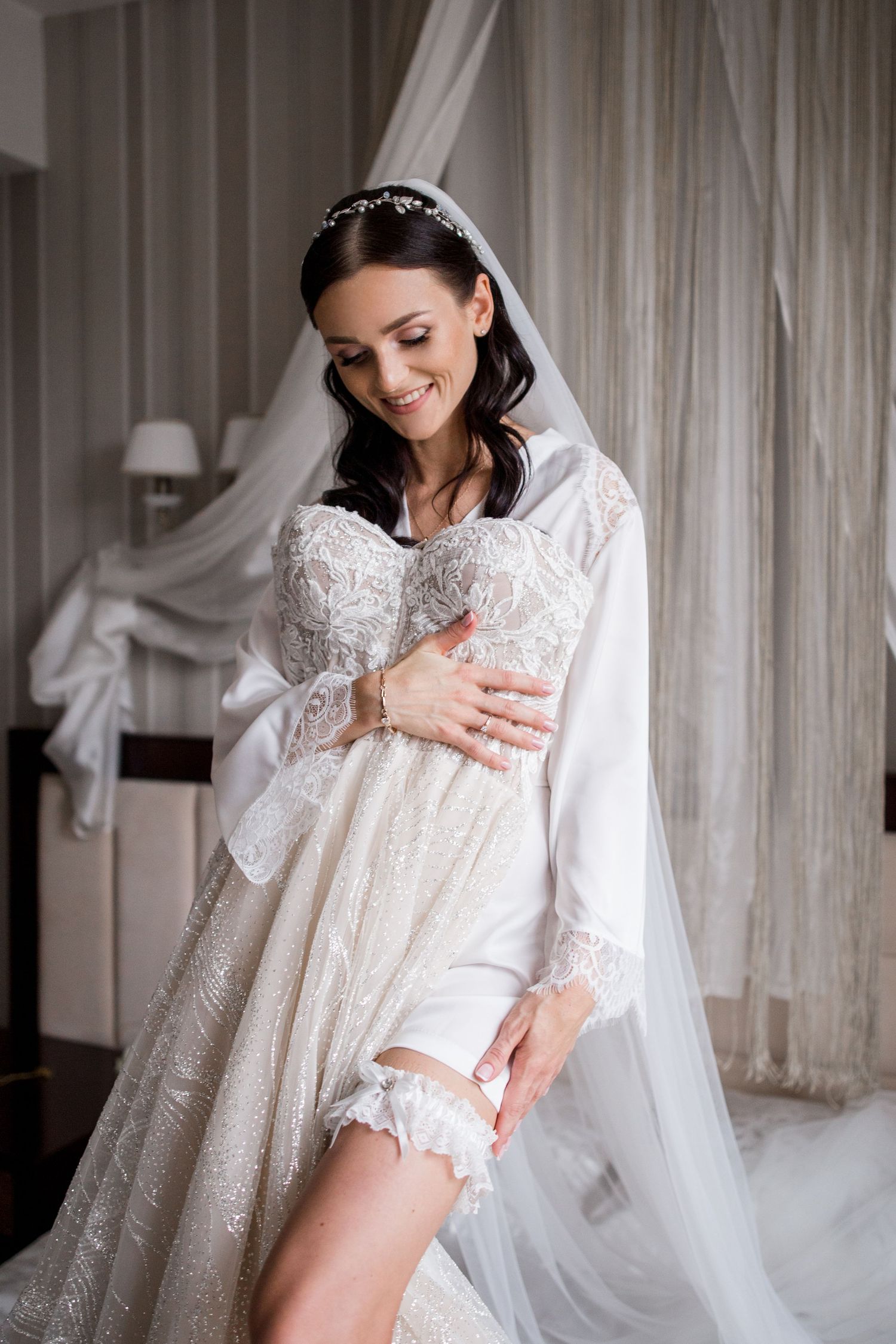 Smiling bride in lace robe and veil adjusting her garter in an elegant white bedroom while holding her wedding dress.