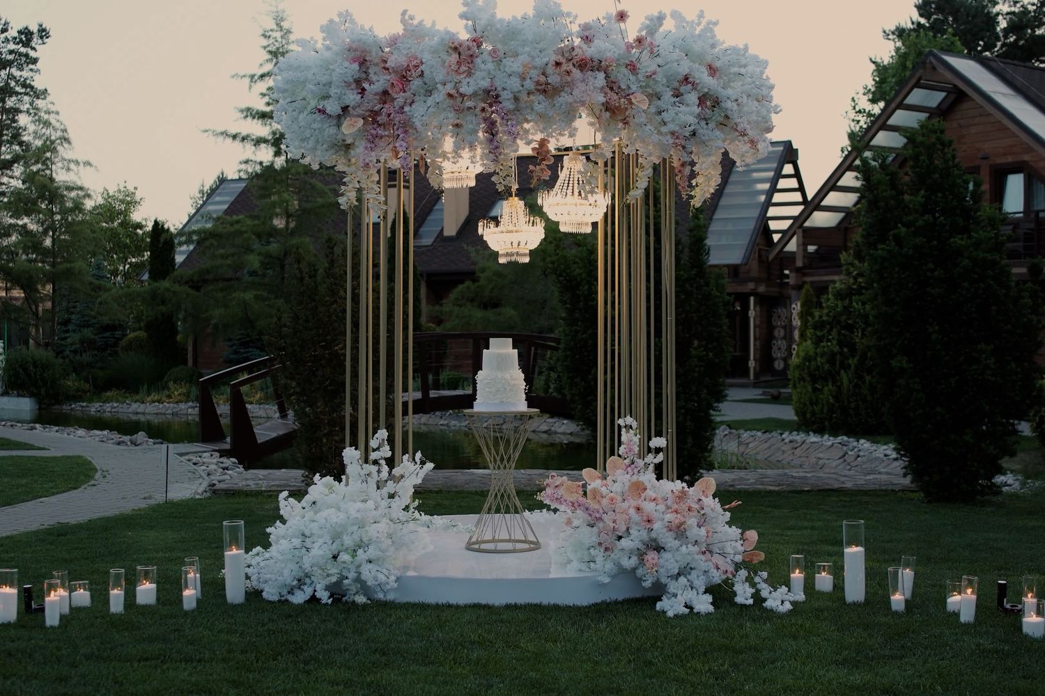 Elegant outdoor wedding cake display with white floral arch, gold candelabras, and candles on a manicured lawn at dusk.