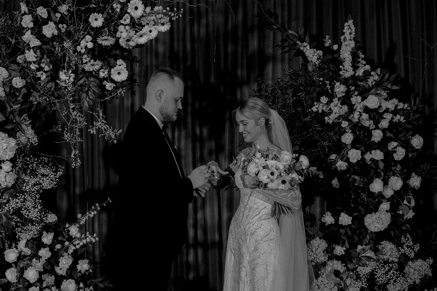 Black and white wedding ceremony photo of bride and groom exchanging rings amid lush floral arrangements.