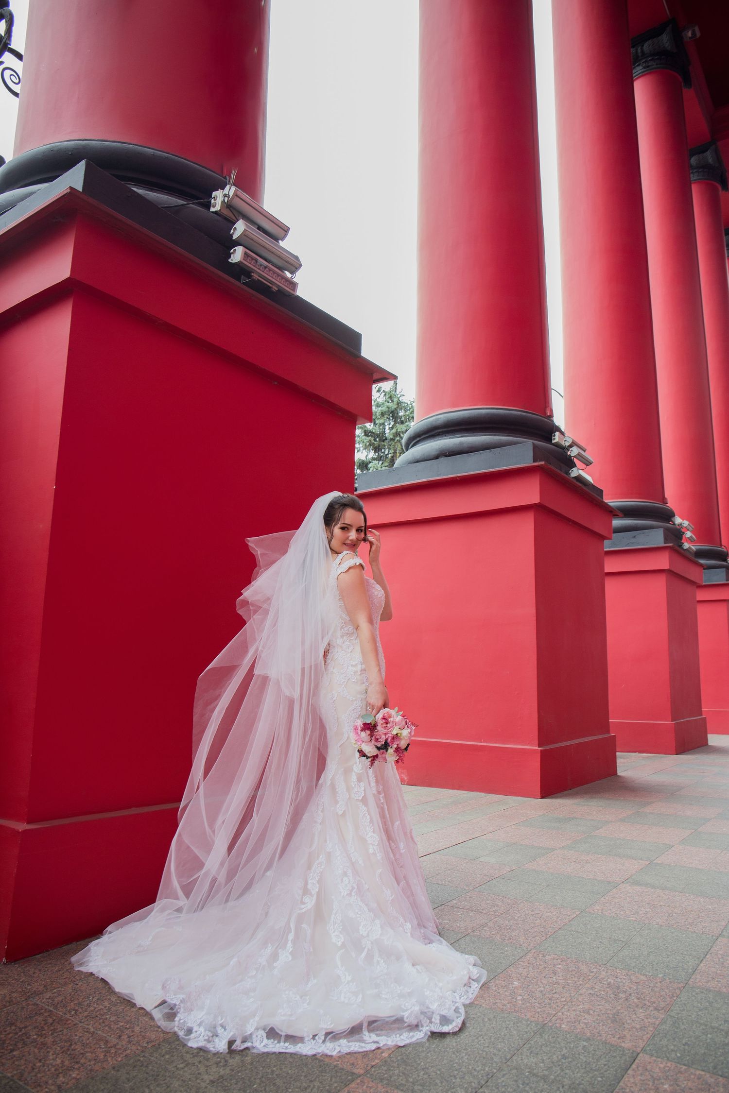 Elegant bride in white lace gown and veil holding bouquet beside tall red columns.