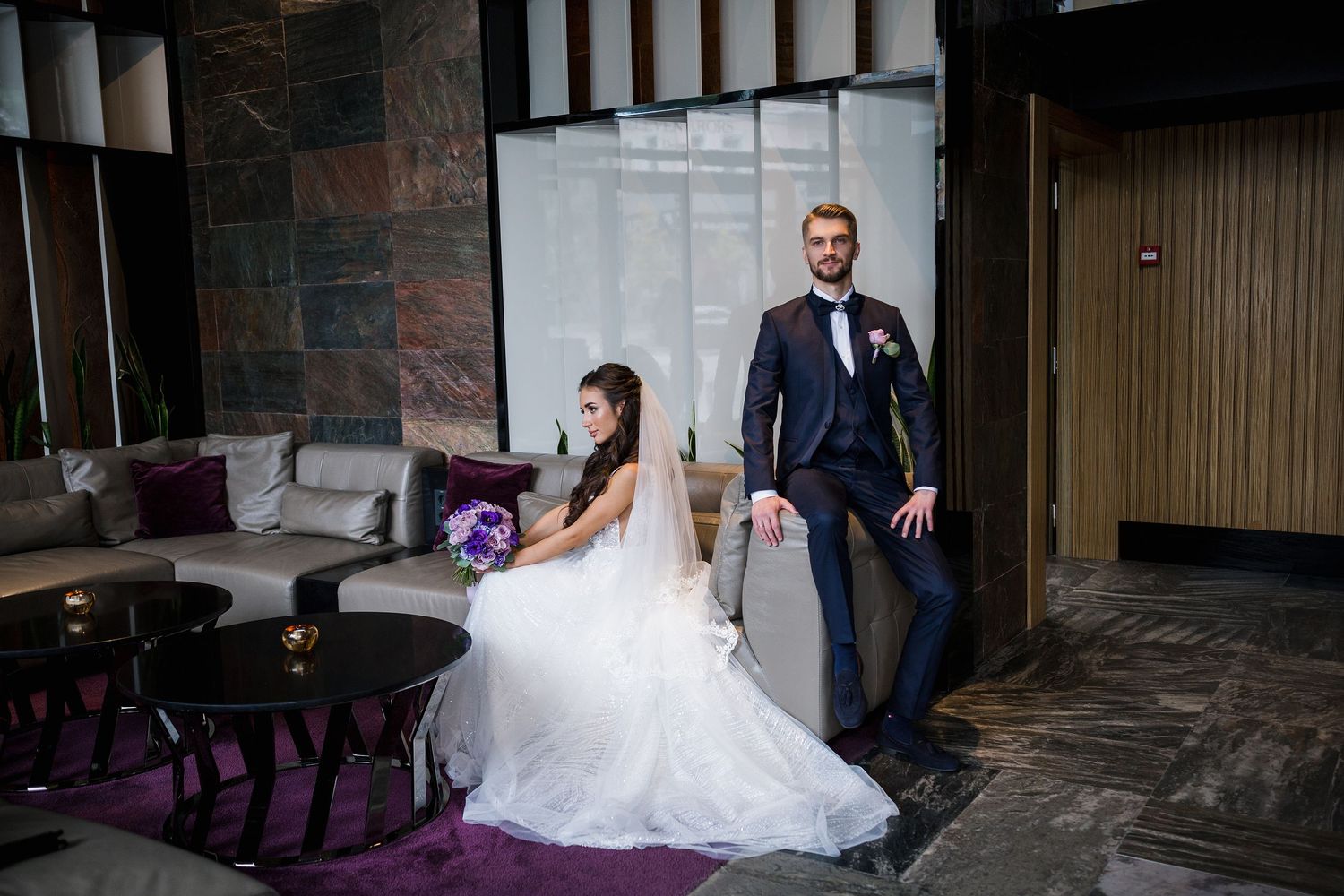 Bride in white gown holding purple bouquet sits beside groom in navy suit in elegant hotel lobby.