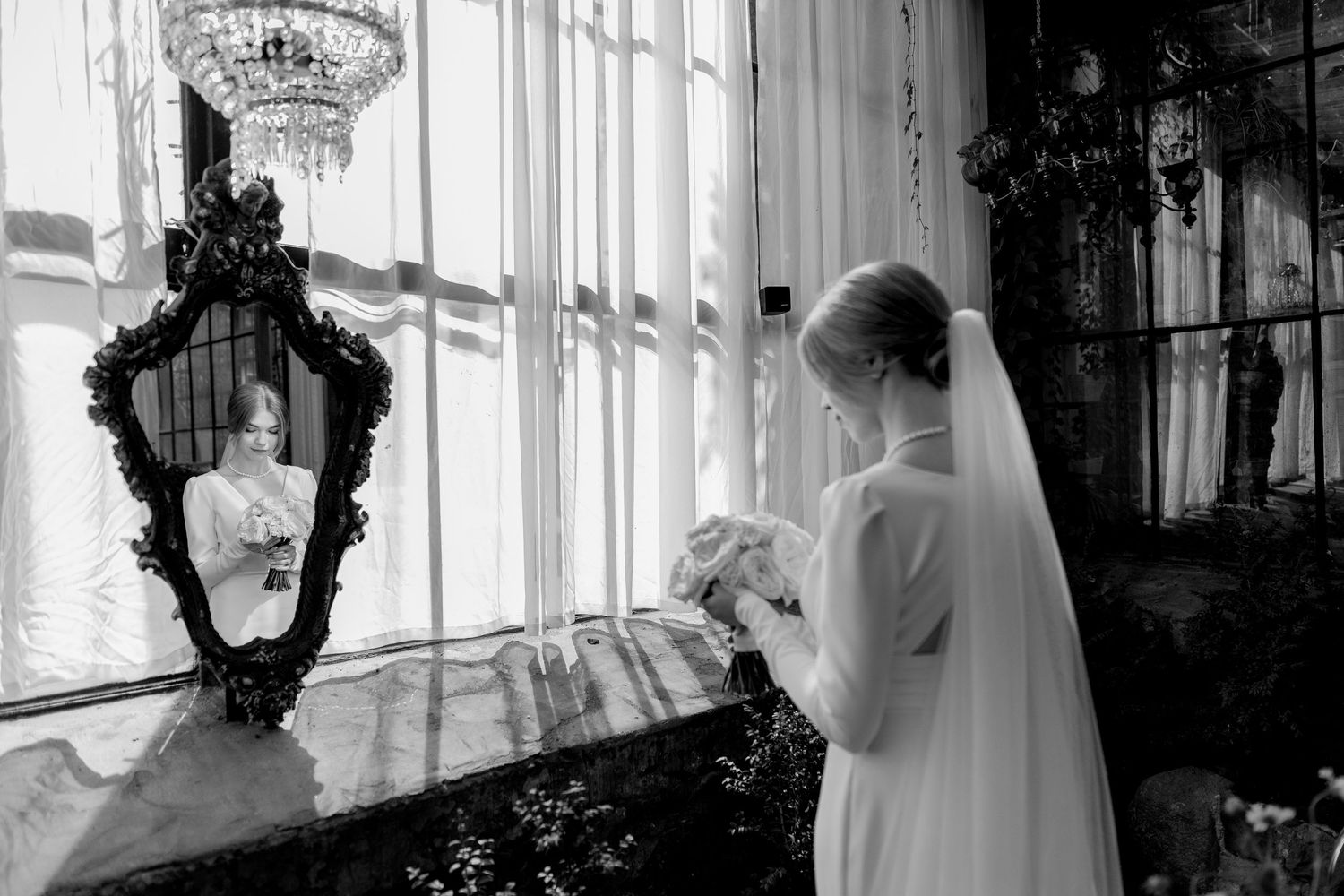 Bride in white gown and veil holds bouquet near ornate mirror reflecting her image in elegant black and white wedding photo.